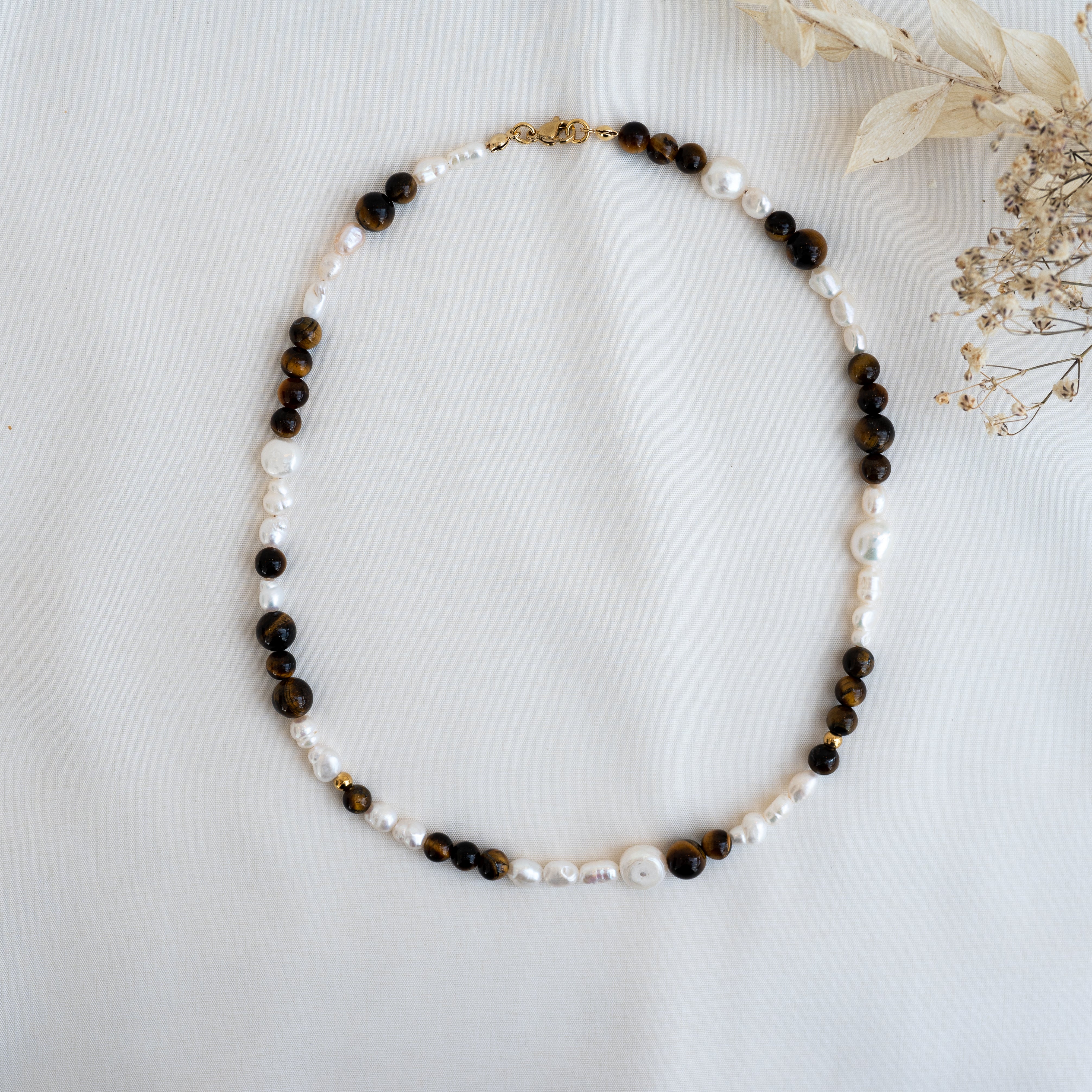Eine Halskette aus abwechselnd weißen Perlen und runden, dunkelbraunen Perlen mit goldenem Verschluss auf weißem Stoff mit getrockneten Blumen in der rechten oberen Ecke.