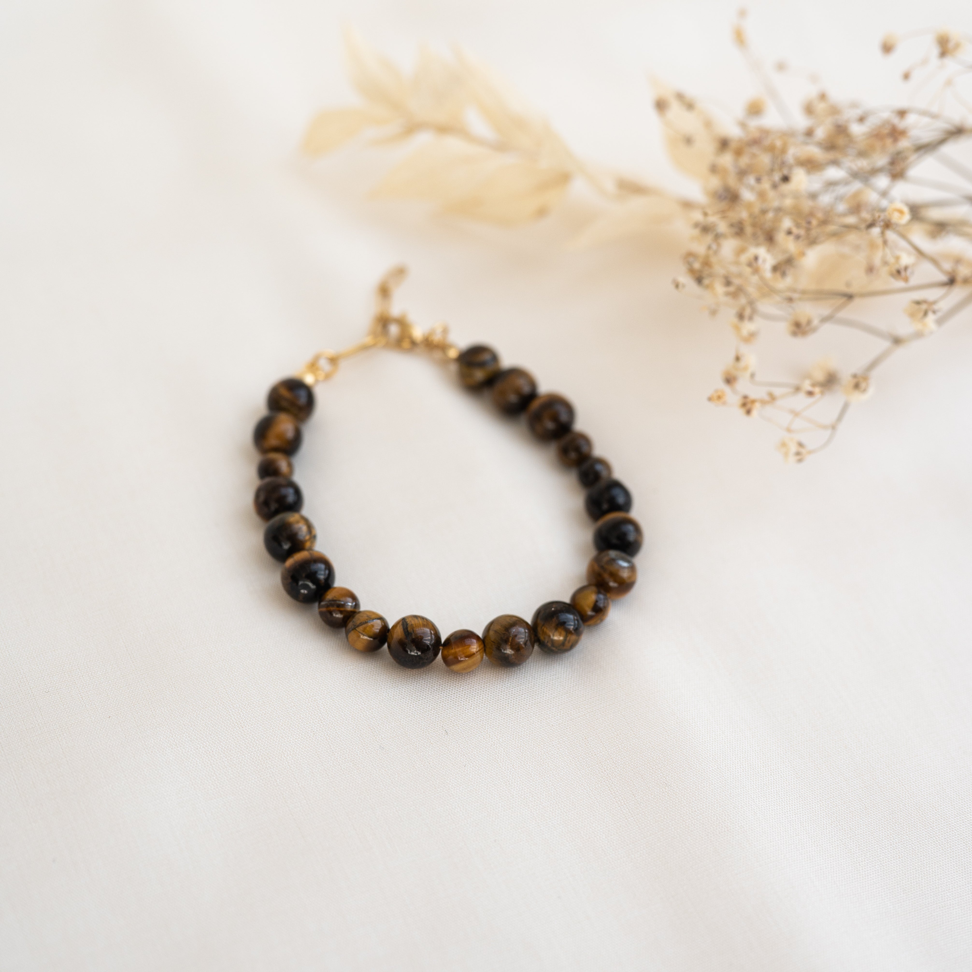 Ein Perlenarmband mit glänzenden braunen und schwarzen Steinen auf einer weißen Oberfläche, daneben getrocknete beige Blätter und zarte Zweige getrockneter Blumen.