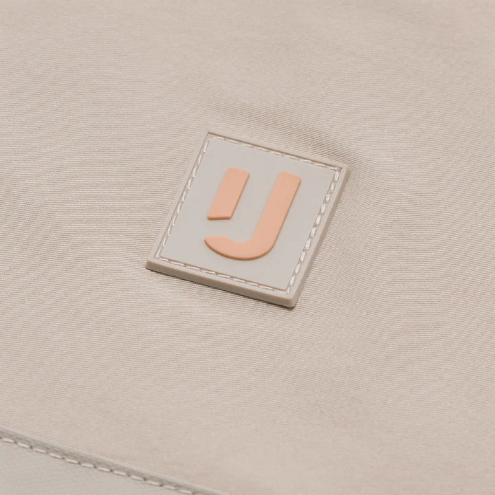 Eine Nahaufnahme eines beigen Stoffaufnähers mit einem erhabenen, stilisierten orangefarbenen J-Logo, das mit weißen Nähten auf einem passenden Hintergrund umrandet ist.
