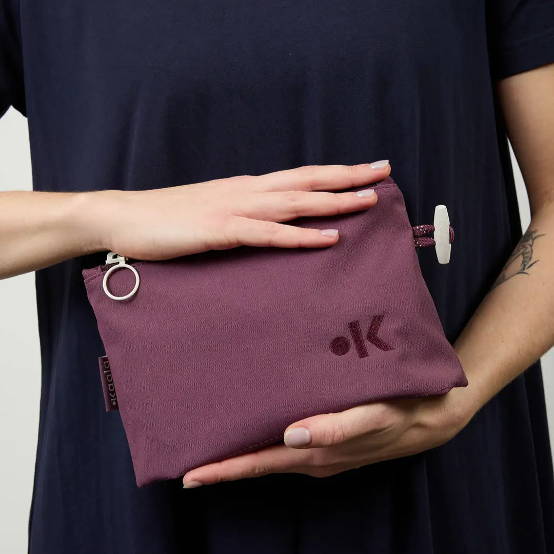 Eine Person, die ein marineblaues Hemd trägt, hält einen lilafarbenen Stoffbeutel mit Reißverschluss, der einen Ringzug und ein kleines Logo auf der Vorderseite aufweist. Das Gesicht der Person ist nicht sichtbar.