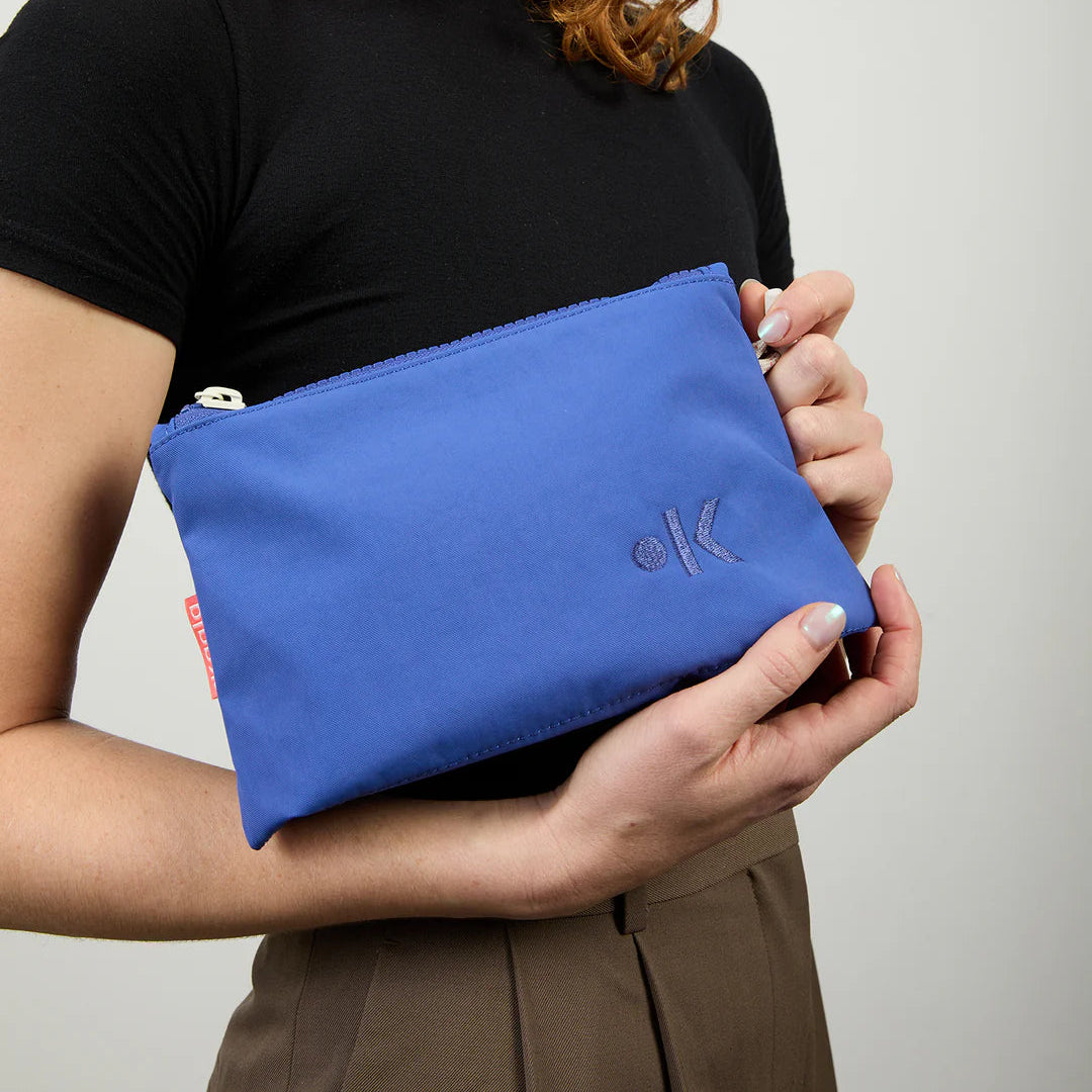 Eine Person mit roten Locken, die ein schwarzes Oberteil und eine braune Hose trägt, hält eine blaue Tasche mit Reißverschluss und einem dezenten Logo auf einer Seite vor einem neutralen Hintergrund.