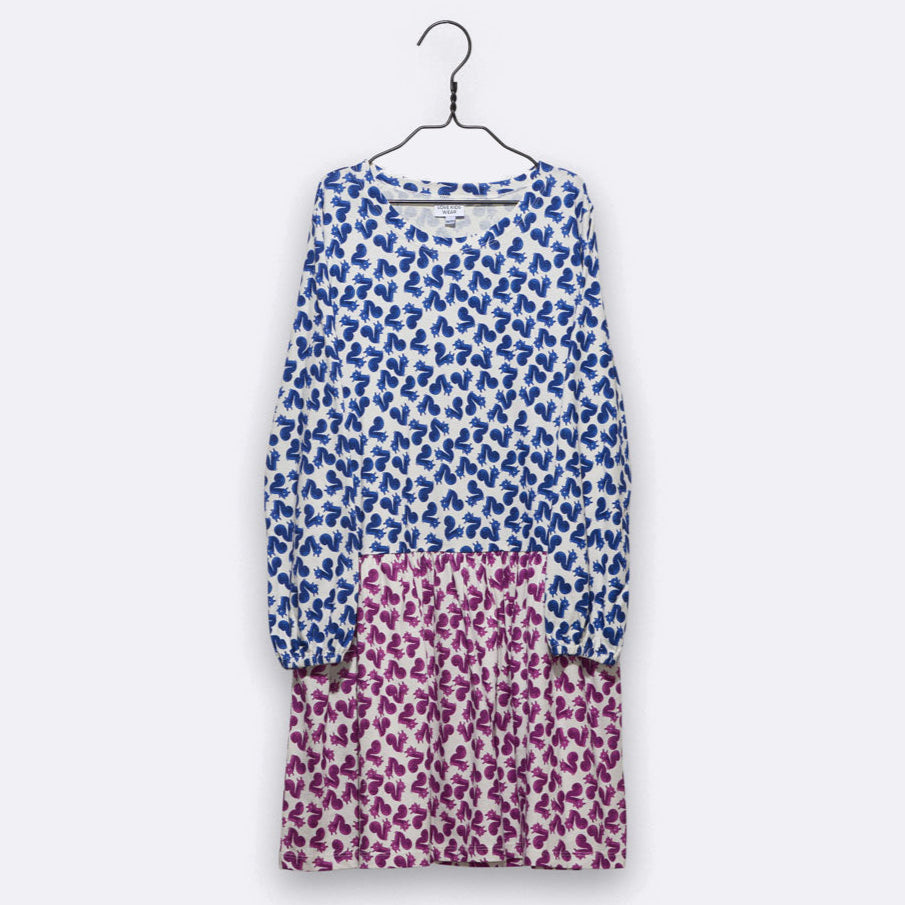 Ein langärmeliges Kleid mit blauem Blumenmuster auf dem Oberteil und den Ärmeln und einem rosafarbenen Blumenmuster auf dem Rock, das auf einem Bügel vor einem schlichten hellen Hintergrund hängt.