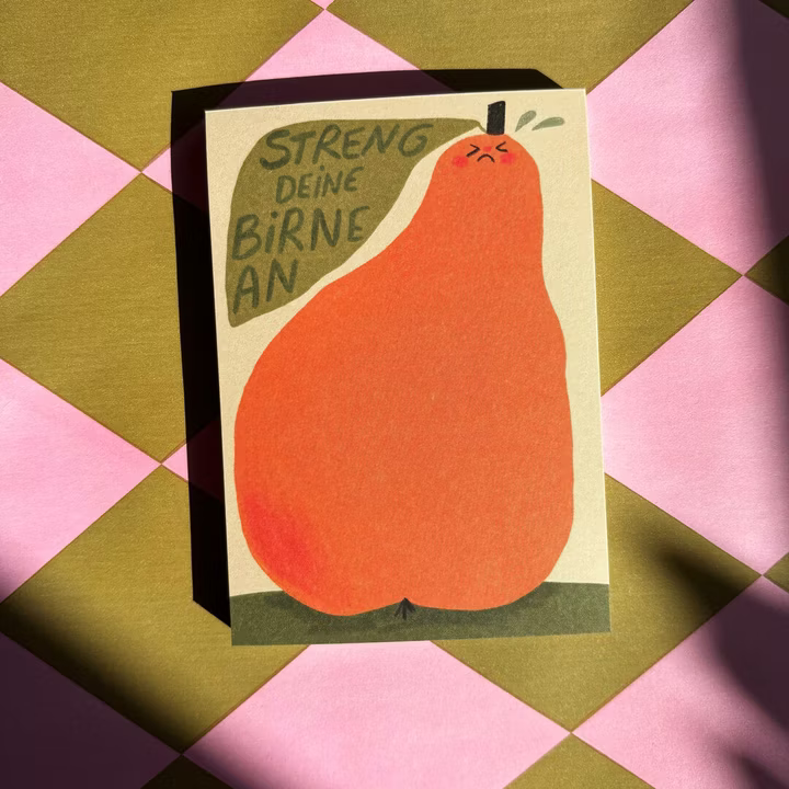 Eine Postkarte mit einer Illustration einer Orangenbirne mit angespanntem Gesicht. Der deutsche Text darüber lautet "Streng deine Birne an". Die Karte befindet sich auf einer rosa und olivfarbenen, rautenförmigen Oberfläche, teilweise im Schatten.