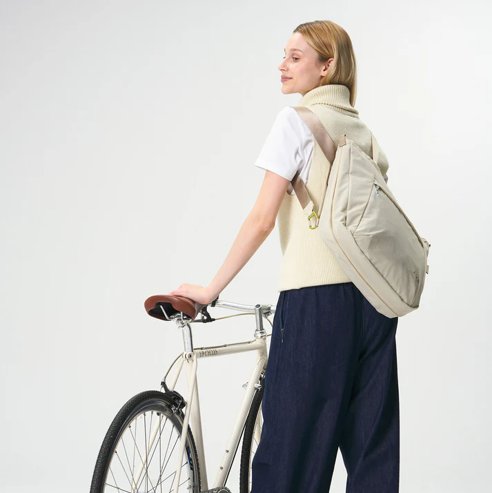 Eine Person, die eine cremefarbene Pulloverweste, ein weißes T-Shirt und eine marineblaue Hose trägt, steht neben einem weißen Fahrrad, lächelt und schaut über ihre Schulter. Sie hat eine beigefarbene Tasche auf dem Rücken.