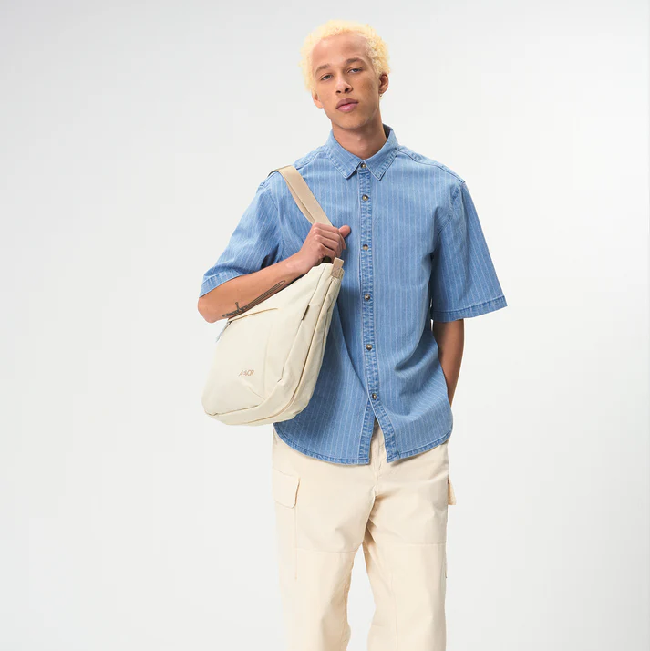 Eine Person mit kurzen, gebleichten blonden Haaren trägt ein blaues, kurzärmeliges Button-up-Hemd und eine cremefarbene Cargohose, hält eine beigefarbene Umhängetasche und steht vor einem schlichten, hellen Hintergrund.