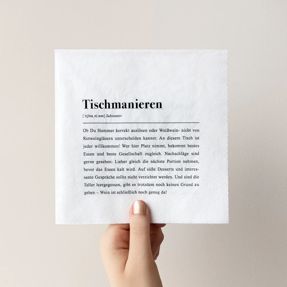 Eine Hand hält eine weiße Serviette hoch, auf der in schwarzer Schrift ein deutscher Text über Tischmanieren vor einem schlichten, hellen Hintergrund steht.