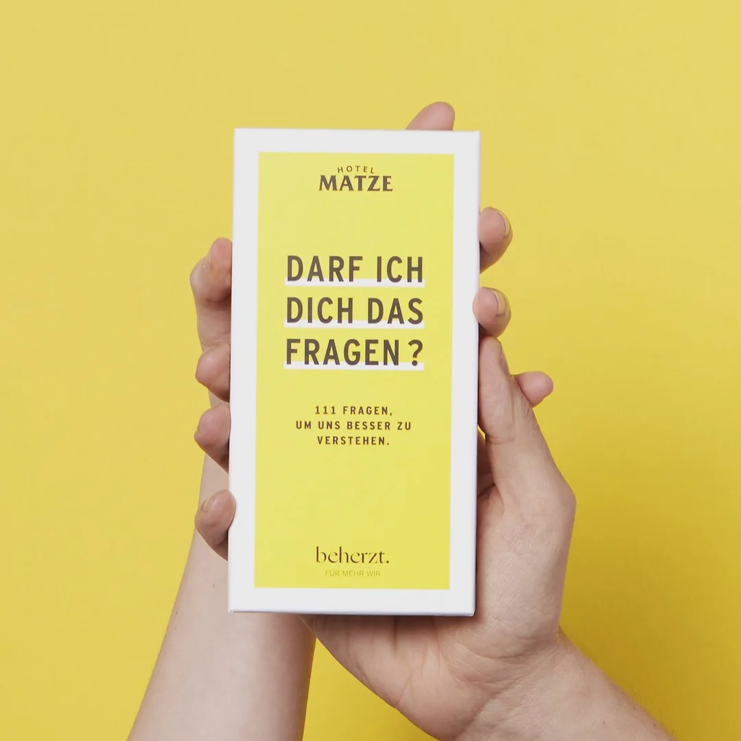 Zwei Hände halten eine gelb-weiße Pappschachtel mit deutscher Aufschrift: Darf ich dich das fragen? 111 Fragen, um uns besser zu verstehen. Der Hintergrund ist einfarbig gelb.