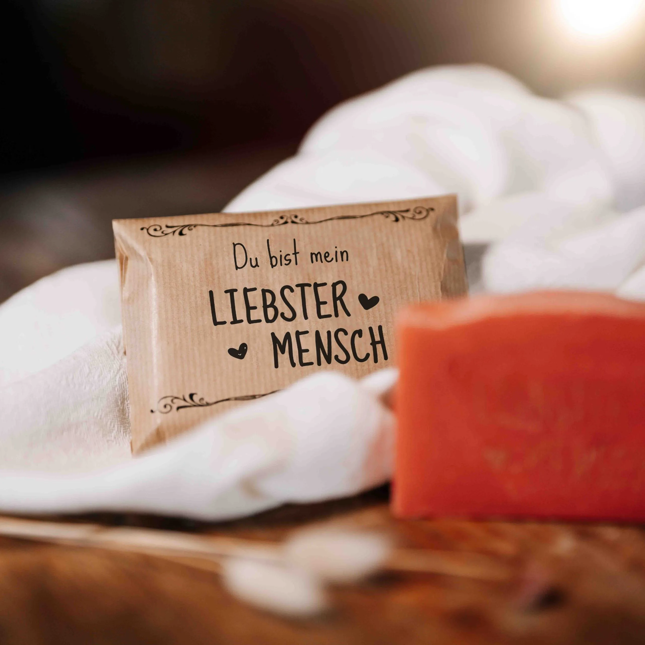 Ein braunes Papierpaket mit dem deutschen Text Du bist mein liebster Mensch liegt auf einem weißen Tuch neben einem orangefarbenen Stück Seife. Der Hintergrund ist sanft verschwommen und warm beleuchtet.