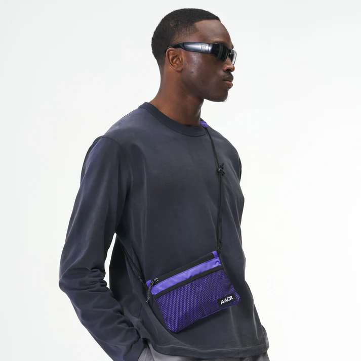 Ein Mann mit einer dunklen Sonnenbrille, einem dunklen langärmeligen Hemd und einer lila Umhängetasche steht vor einem schlichten weißen Hintergrund.