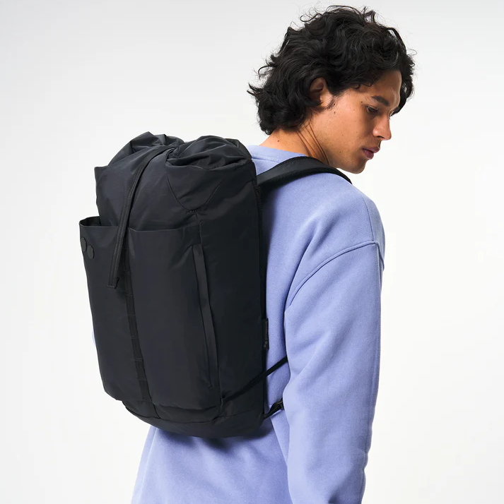 Eine Person, die ein hellviolettes Sweatshirt trägt, hat einen großen schwarzen Rucksack auf dem Rücken und blickt über ihre Schulter auf einen schlichten weißen Hintergrund.