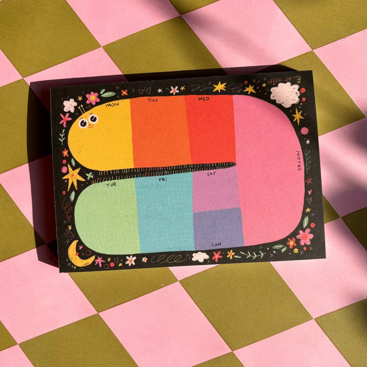 Ein farbenfroher Wochenplaner mit Abschnitten für jeden Tag auf einem schwarzen Hintergrund, der mit Blumen, Wolken und Sternen verziert ist. Der Planer ist auf einer rosa und olivgrün karierten Oberfläche platziert.