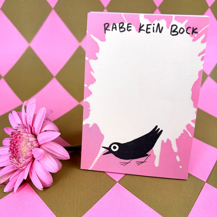 Eine rosafarbene Notizkarte mit einem schwarzen Cartoon-Vogel und weißen Farbspritzern mit der Aufschrift RABE KEIN BOCK steht neben einer rosafarbenen Gänseblümchenblüte auf einem rosa-gold-karierten Hintergrund.