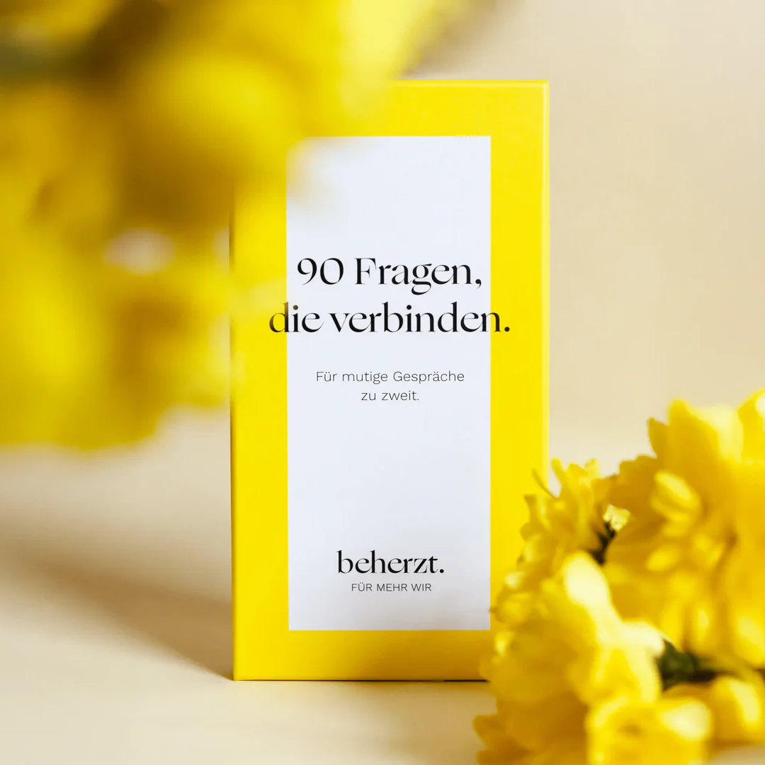 Eine gelbe Schachtel mit dem Titel 90 Fragen, die verbinden steht aufrecht, umgeben von verschwommenen gelben Blumen. Die Schachtel enthält einen Text in deutscher Sprache und ein weißes rechteckiges Etikett auf der Vorderseite.