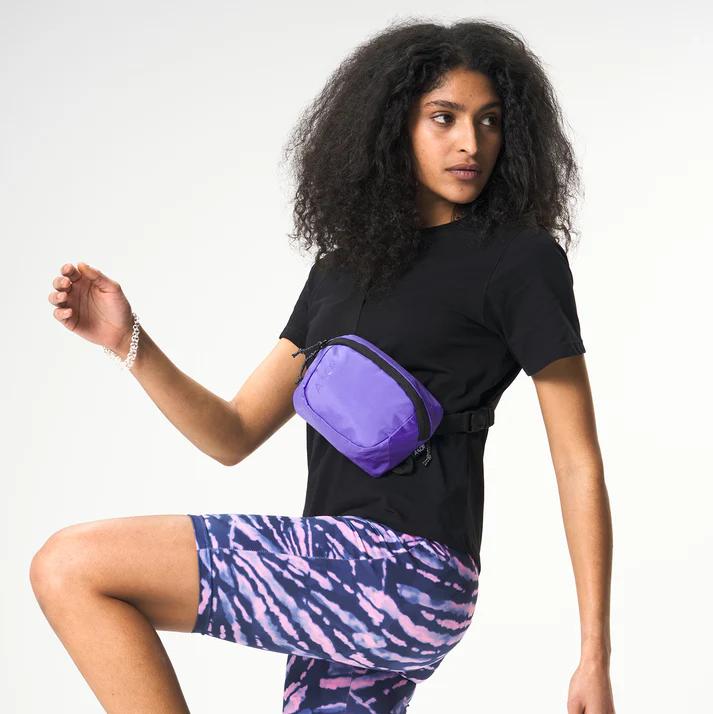 Eine Person mit lockigem Haar posiert selbstbewusst in einem schwarzen T-Shirt, lila gemusterten Shorts und einer lila Hüfttasche, hebt ein Knie und blickt zur Seite vor einem schlichten Hintergrund.