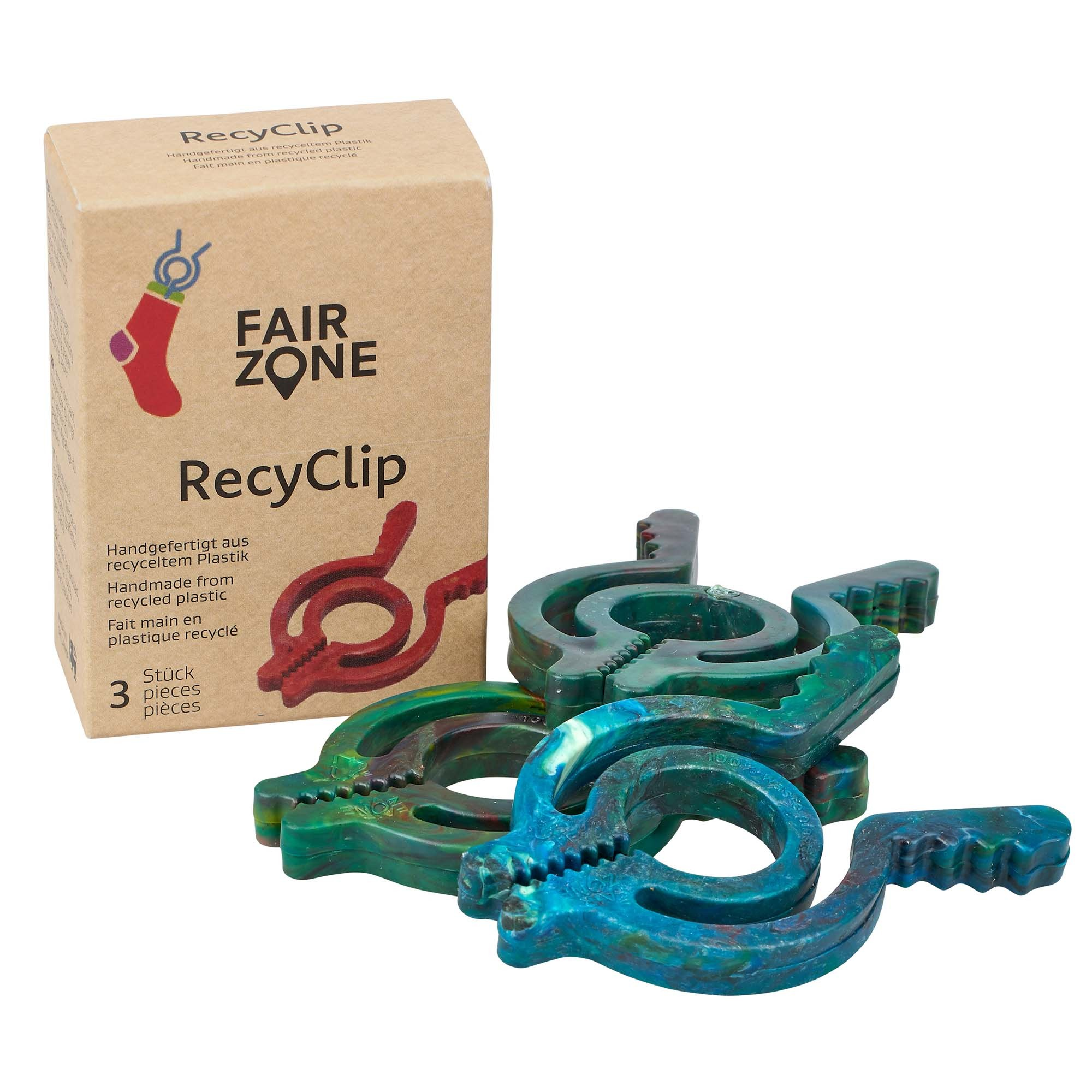 Ein Karton mit der Aufschrift FAIR ZONE RecyClip steht neben drei blau-grünen, wie Wäscheklammern geformten Clips aus recyceltem Kunststoff. Der Karton zeigt an, dass sich darin drei Stück befinden, die aus recyceltem Kunststoff handgefertigt sind.