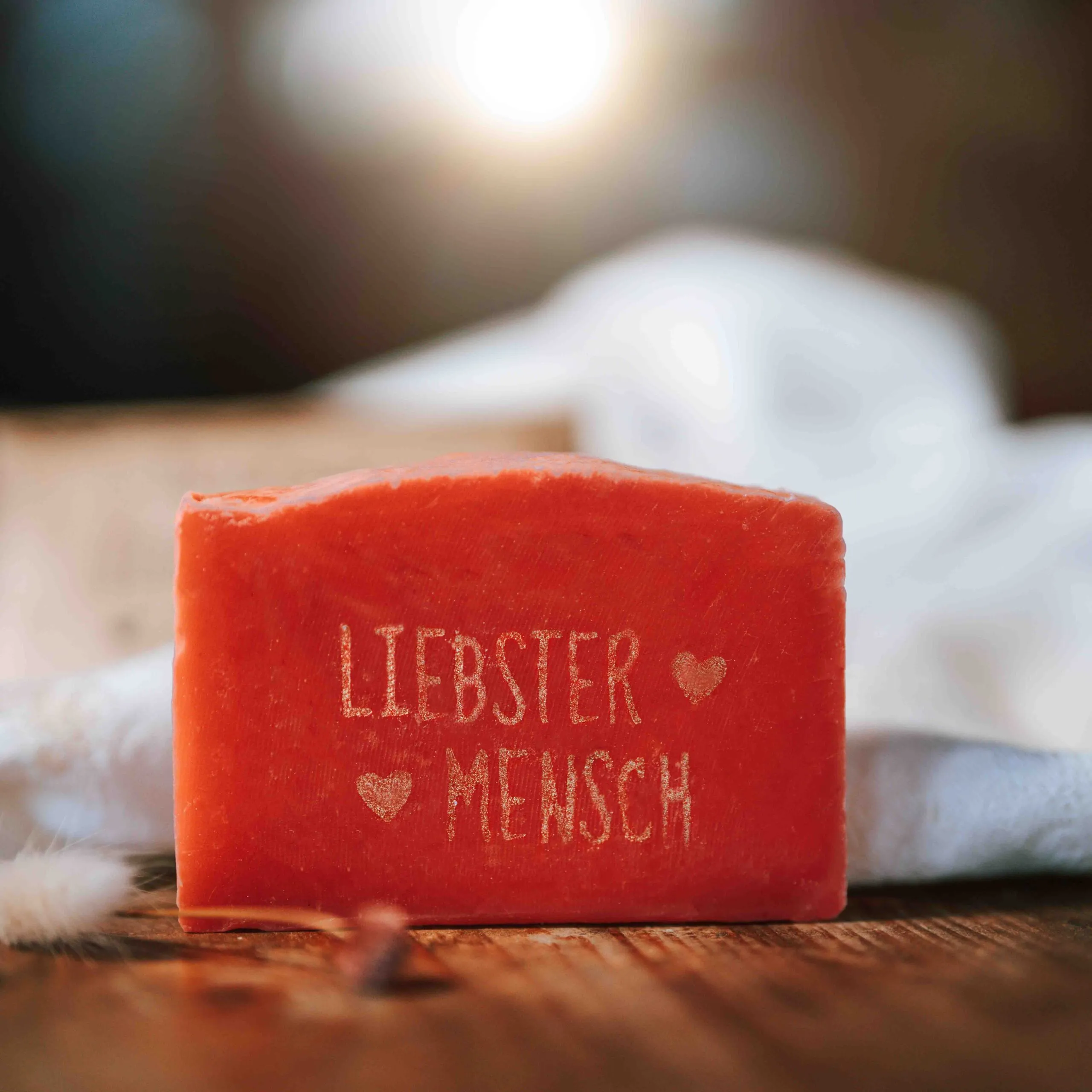 Ein rötlich-oranges Seifenstück mit der Aufschrift LIEBSTER MENSCH und zwei kleinen eingravierten Herzen, platziert auf einem weichen weißen Tuch, mit einem warmen, sanft verschwommenen Hintergrund.