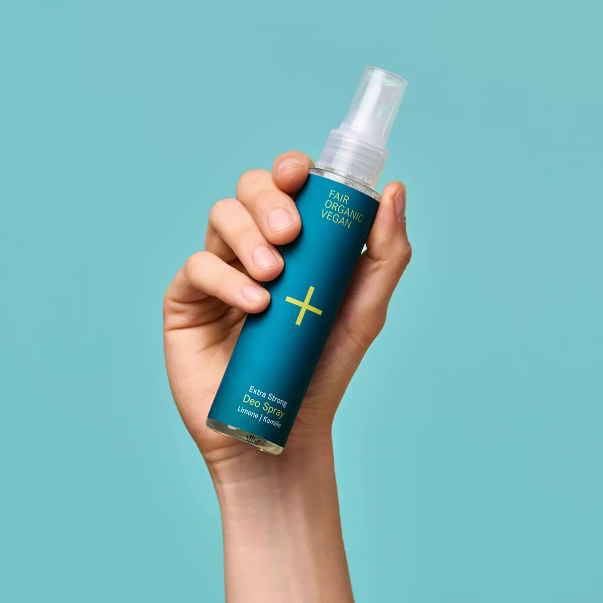 Eine Hand hält eine blaue Sprühflasche mit der Aufschrift Fair Organic Vegan Extra Strong Deo Spray vor einem schlichten türkisfarbenen Hintergrund.