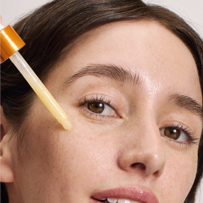 Eine Nahaufnahme des Gesichts einer Person mit klarer Haut, die einen Tropfen eines gelblichen Serums in der Nähe ihrer Wange hält und sich darauf vorbereitet, ein Hautpflegeprodukt aufzutragen.