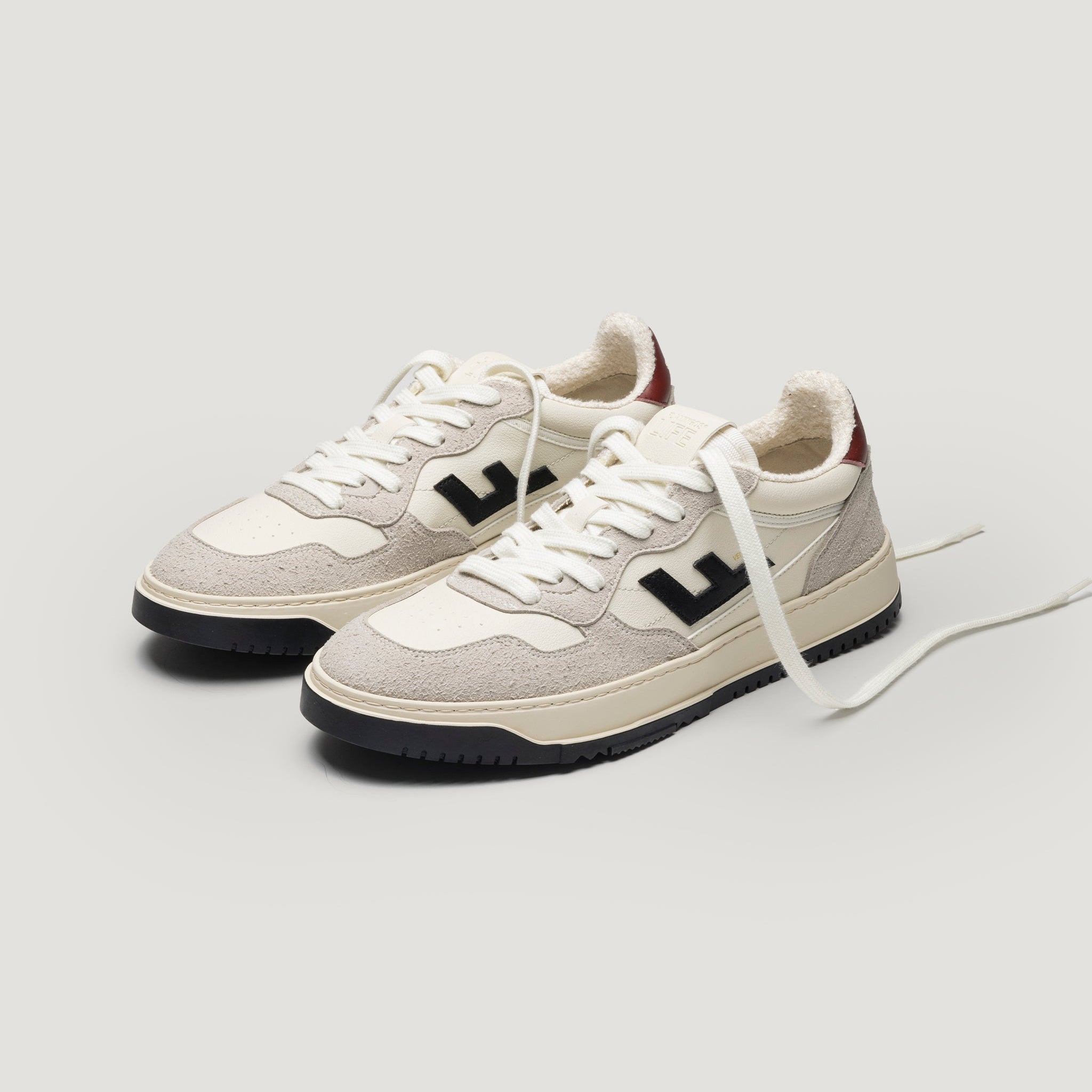 Ein Paar cremefarbene und beigefarbene Turnschuhe mit schwarzen Akzenten, schwarzer Sohle und weißen Schnürsenkeln, mit einem großen schwarzen "L"-Logo an der Seite und einem Hauch von Rot an der Ferse, auf einem schlichten hellen Hintergrund.