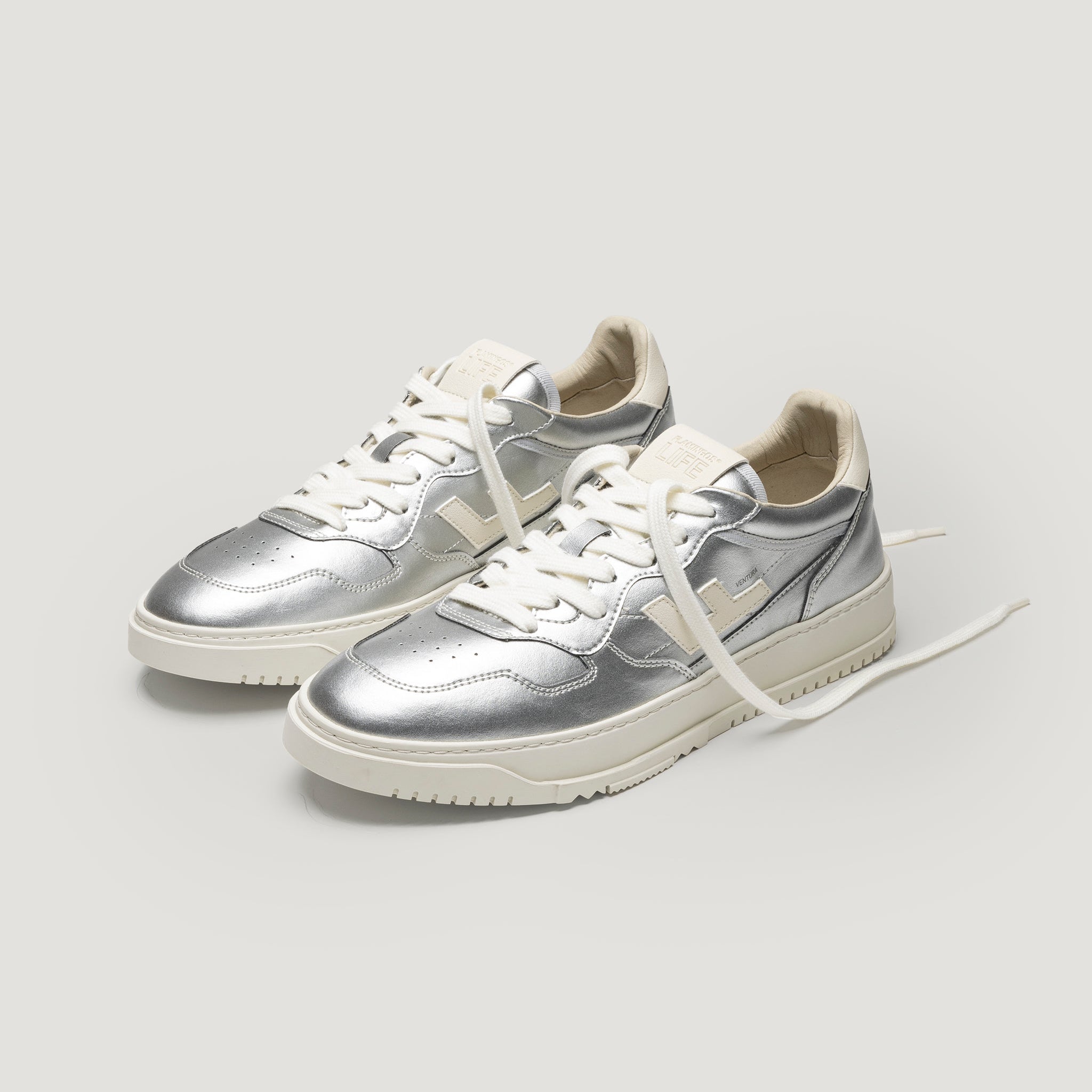 Ein Paar silberfarbene Low-Top-Sneakers mit weißen Schnürsenkeln und cremefarbenen Sohlen vor einem schlichten hellen Hintergrund.