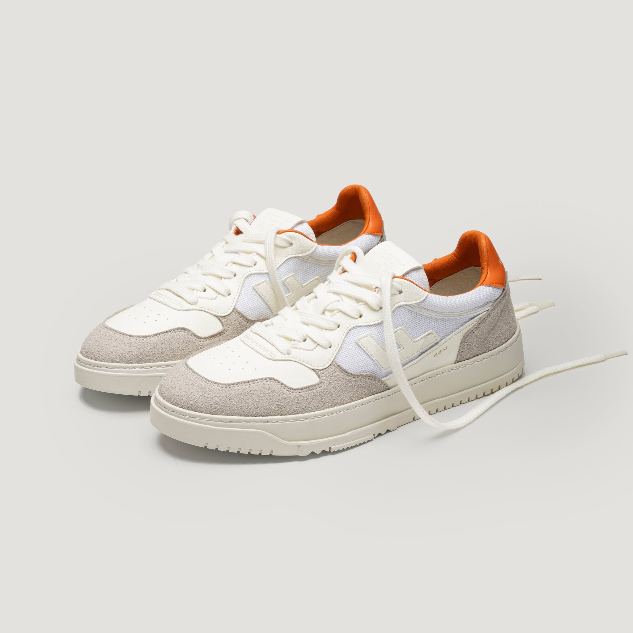 Ein Paar weiße und hellgraue Turnschuhe mit orangefarbenen Akzenten an der Ferse, weißen Schnürsenkeln und strukturierten Sohlen, dargestellt auf einem schlichten hellen Hintergrund.