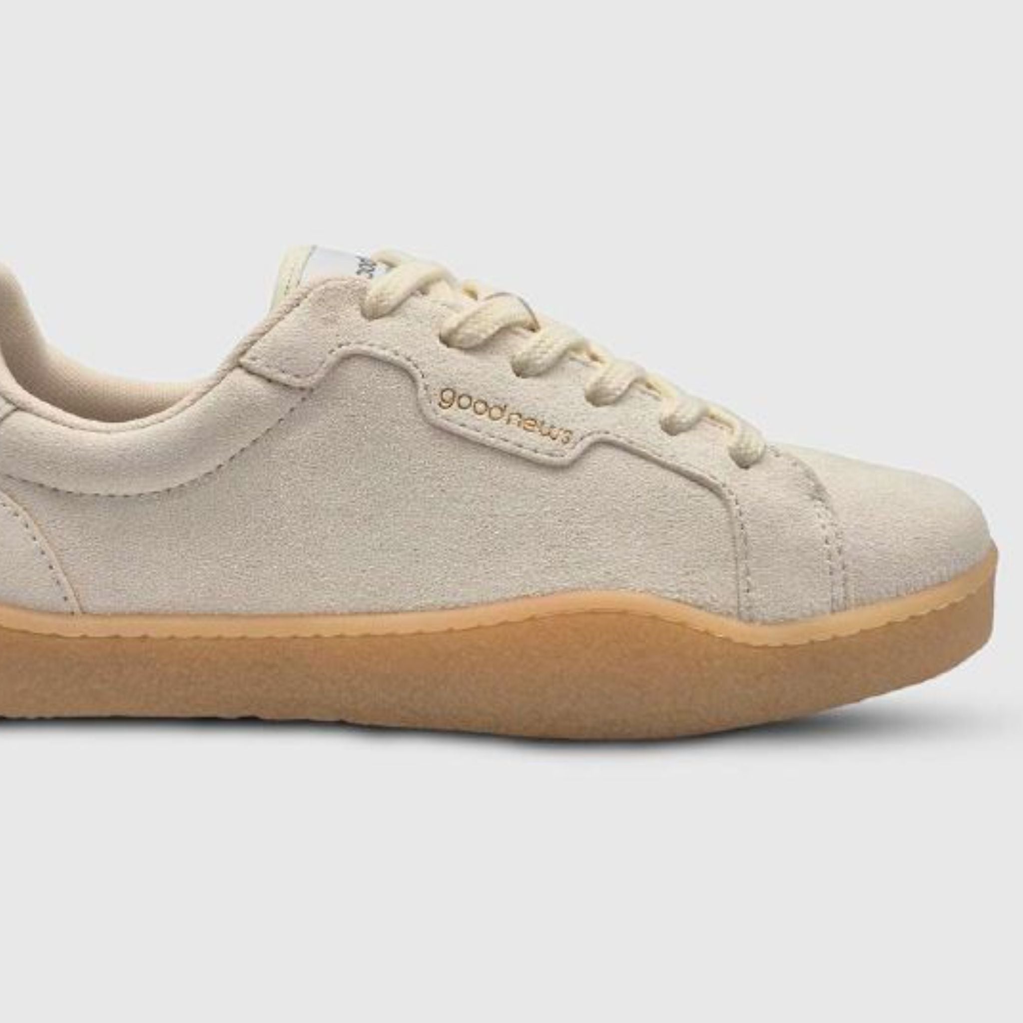 Ein beiger Wildleder-Sneaker mit hellbrauner Gummisohle, beigen Schnürsenkeln und dem Wort "Good News" an der Seite, das auf einem einfarbigen hellgrauen Hintergrund eingeprägt ist.