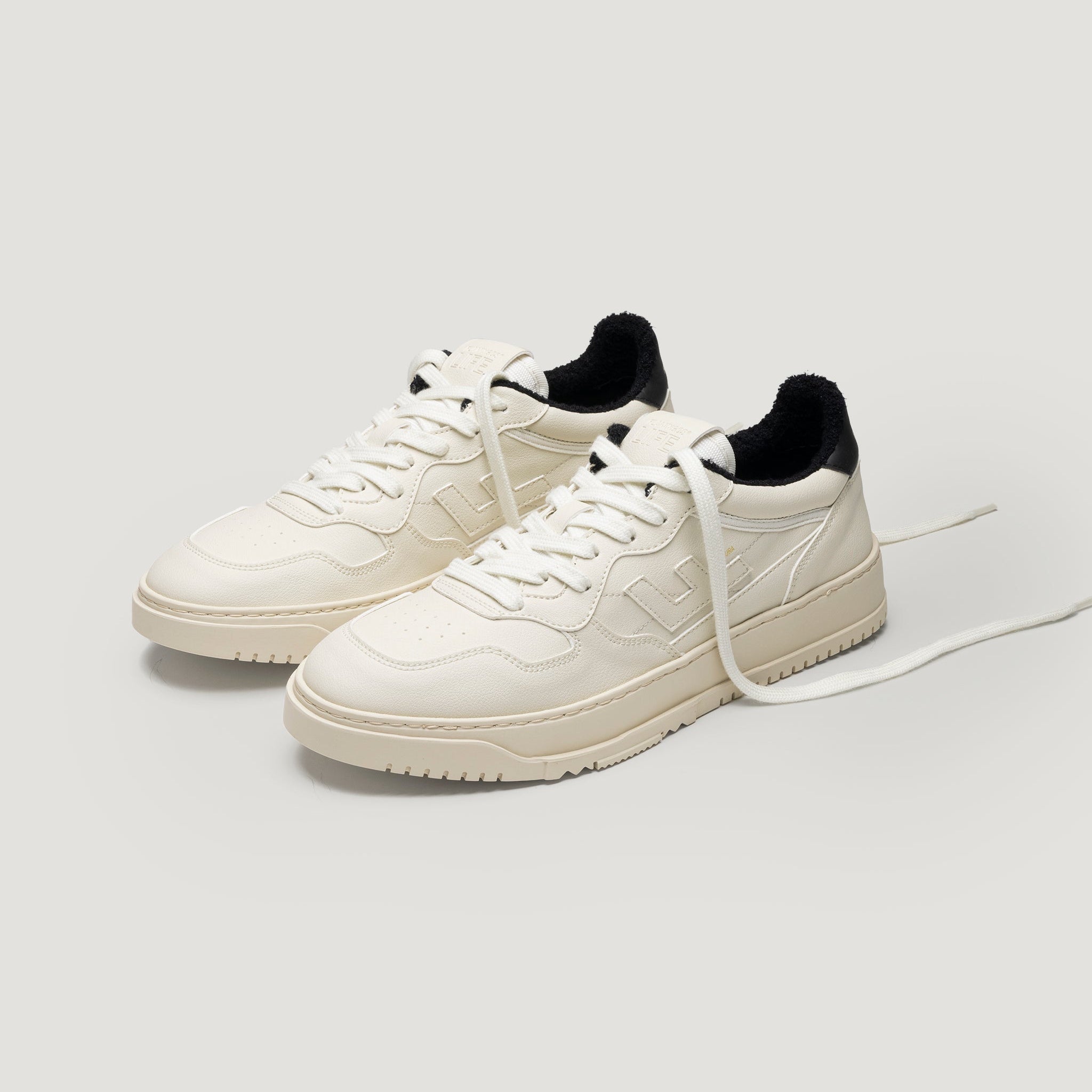 Ein Paar cremefarbene Turnschuhe mit schwarzem Innenleben, weißen Schnürsenkeln und strukturierten Sohlen sind auf einem hellgrauen Hintergrund abgebildet. Ein Schnürsenkel ist locker nach außen verlängert.