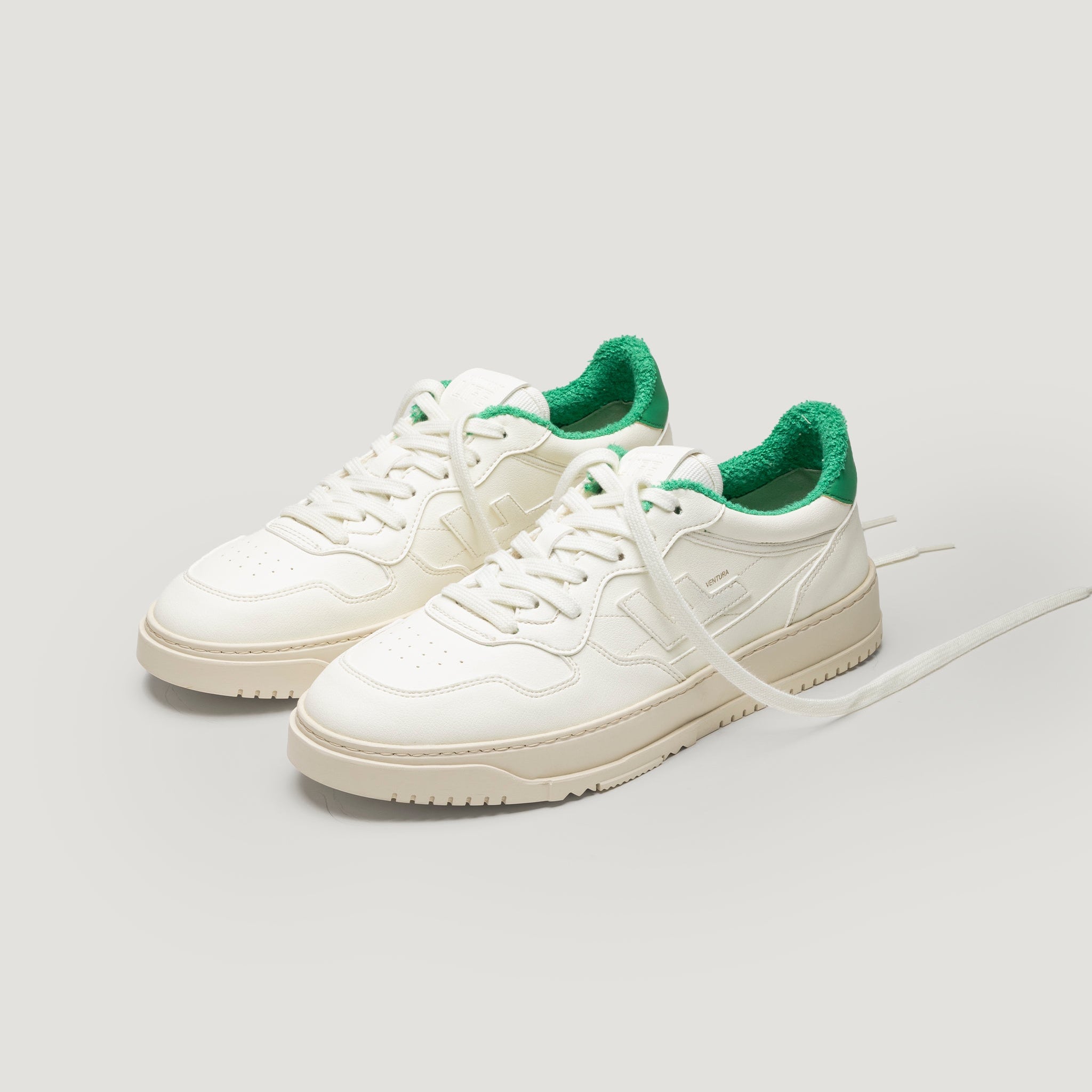 Ein Paar weiße Turnschuhe mit beiger Sohle und grün gepolsterter Innenseite sind nebeneinander auf einem hellen Hintergrund platziert, wobei die Schnürsenkel locker gelöst sind und ein Schnürsenkel nach außen ragt.