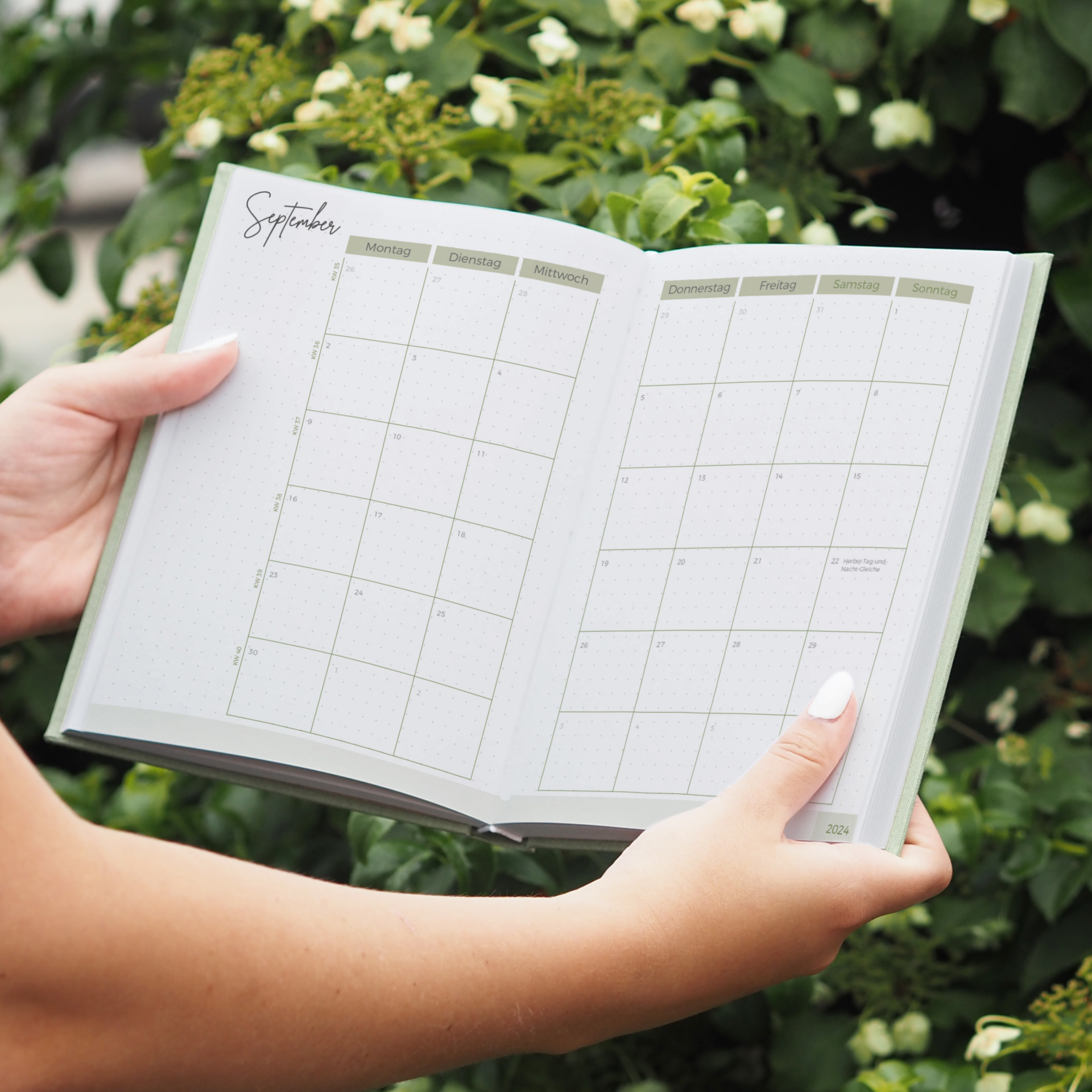 Person hält einen offenen Kalender für September vor einem grünen Hintergrund mit Blumen.