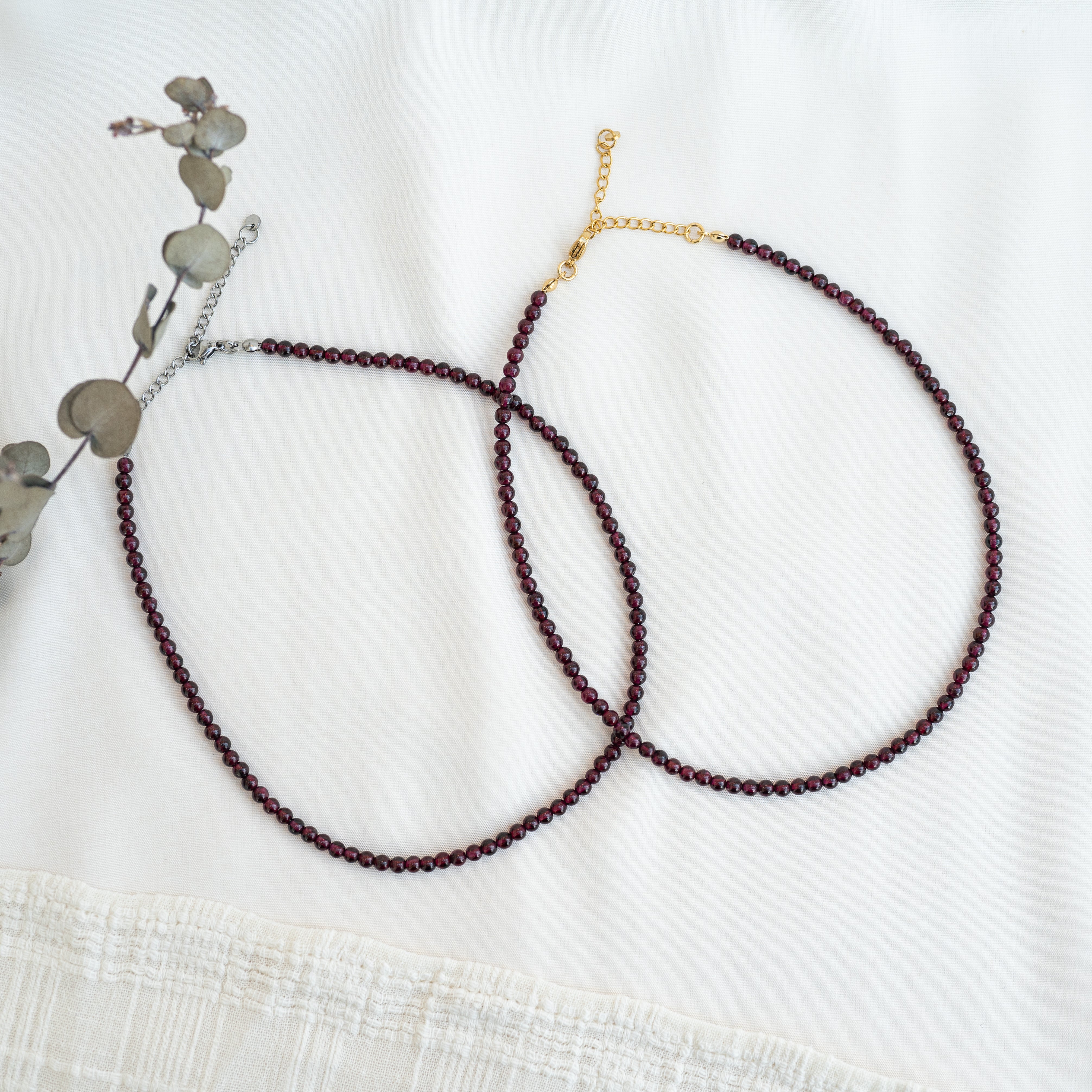 Zwei zarte Perlenketten mit dunkelroten Perlen sind in sich überschneidenden Kreisen auf weißem Stoffgrund angeordnet. Eine Kette hat einen goldenen Verschluss und eine Kette, die andere einen silbernen Verschluss. Auf der linken Seite ist teilweise eine getrocknete Pflanze zu sehen.