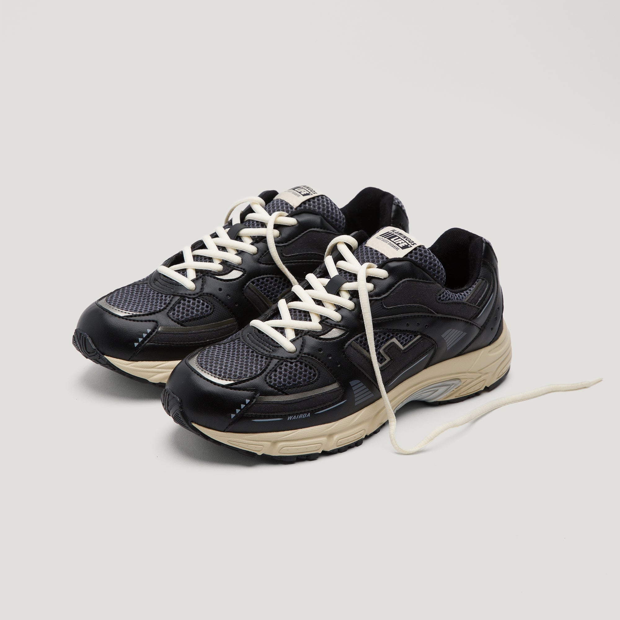 Ein Paar schwarzer und dunkelgrauer Turnschuhe mit beiger Sohle und weißen Schnürsenkeln auf einem schlichten hellen Hintergrund; ein Schnürsenkel ist locker nach vorne verlängert.