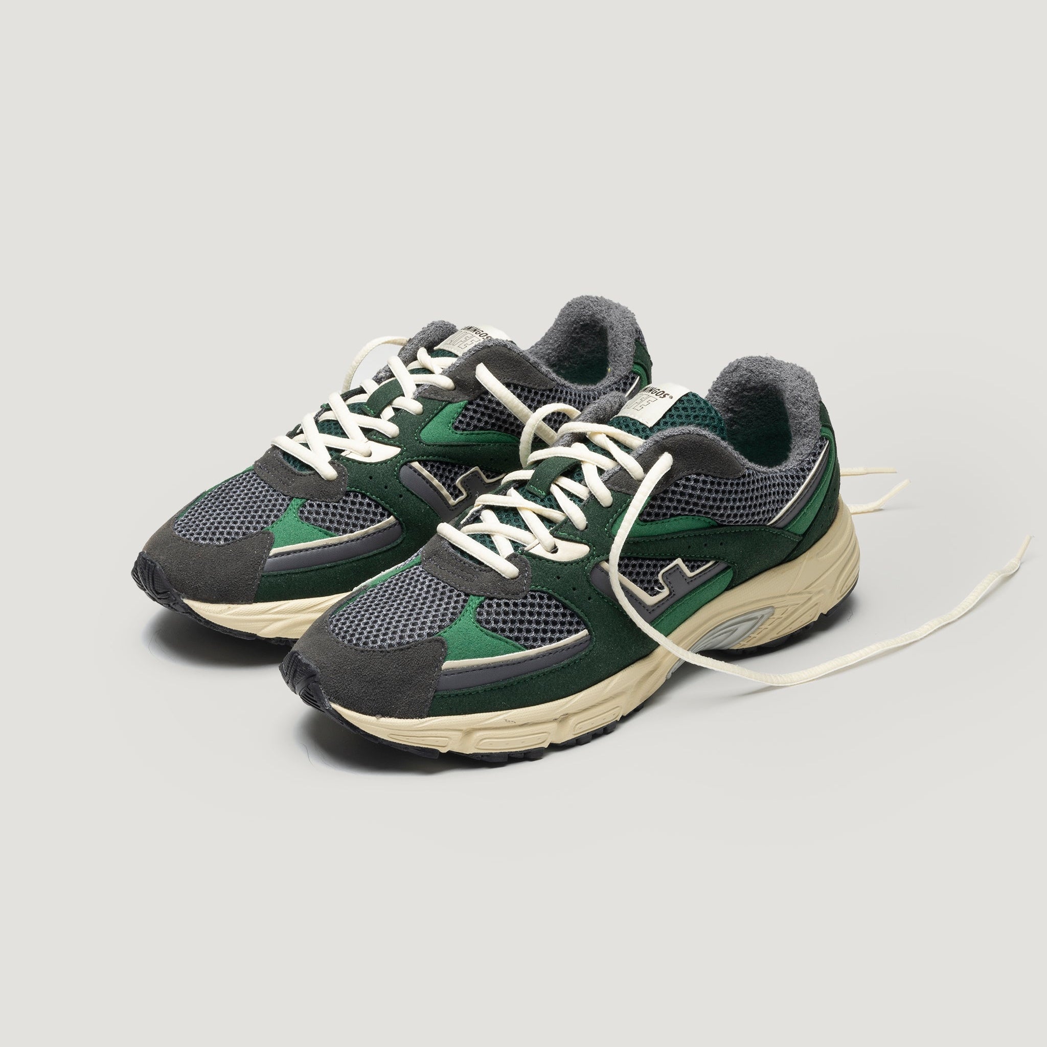 Ein Paar grüne und schwarze Sportschuhe mit weißen Schnürsenkeln und beigen Sohlen, nebeneinander auf einem schlichten hellgrauen Hintergrund.