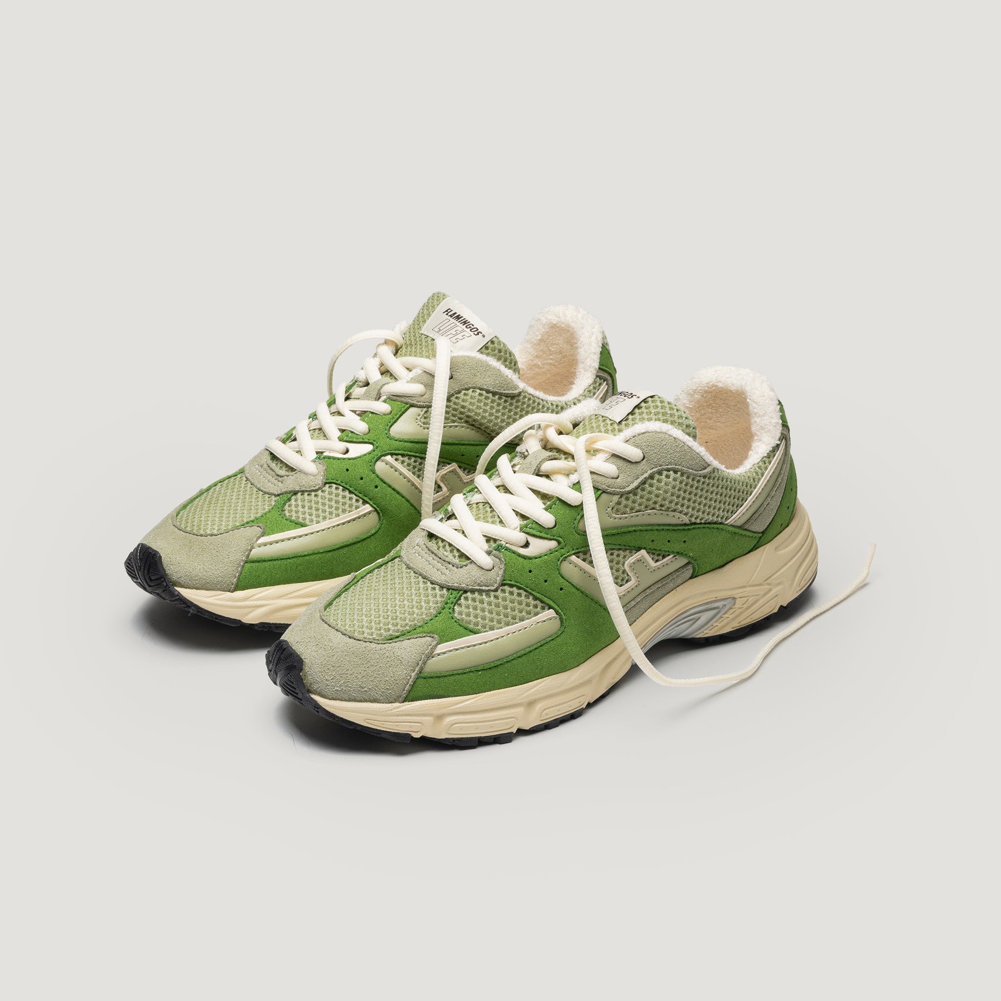 Ein Paar grüne und beigefarbene Sneakers mit weißen Schnürsenkeln und schwarzen Sohlen, die vor einem schlichten, hellen Hintergrund präsentiert werden. Die Sneaker haben ein Retro-Design und sichtbare Details aus Mesh und Wildleder.