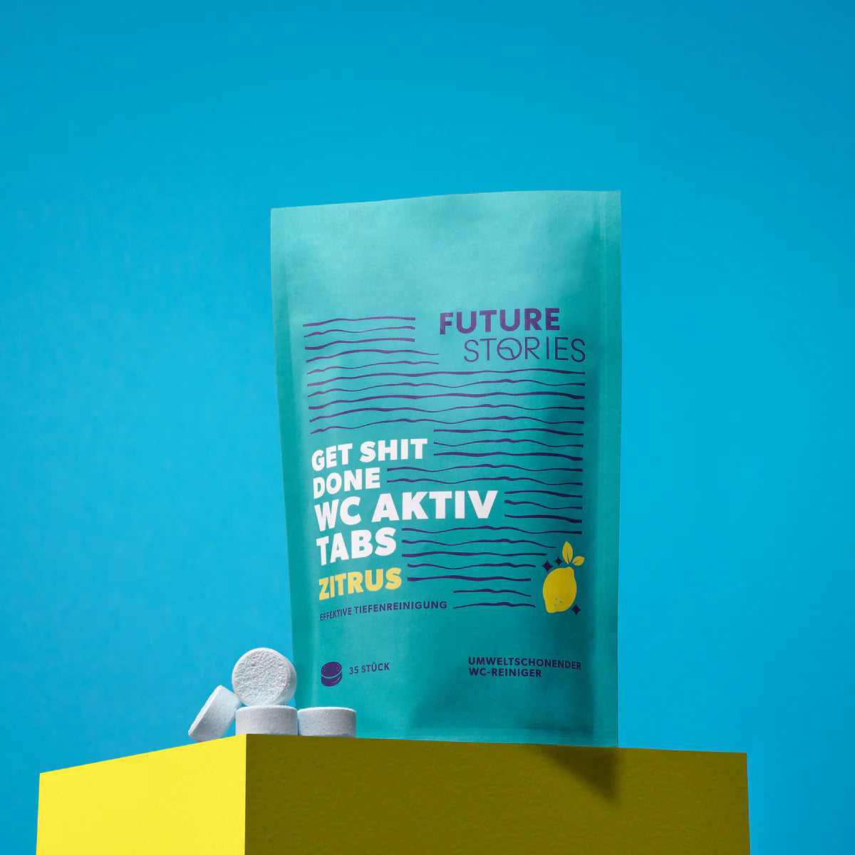 Ein türkisfarbener Beutel mit der Aufschrift Future Stories Get Shit Done WC Aktiv Tabs Zitrus steht aufrecht auf einem gelben Block. In der Nähe liegen mehrere weiße Reinigungstabletten vor einem hellblauen Hintergrund.