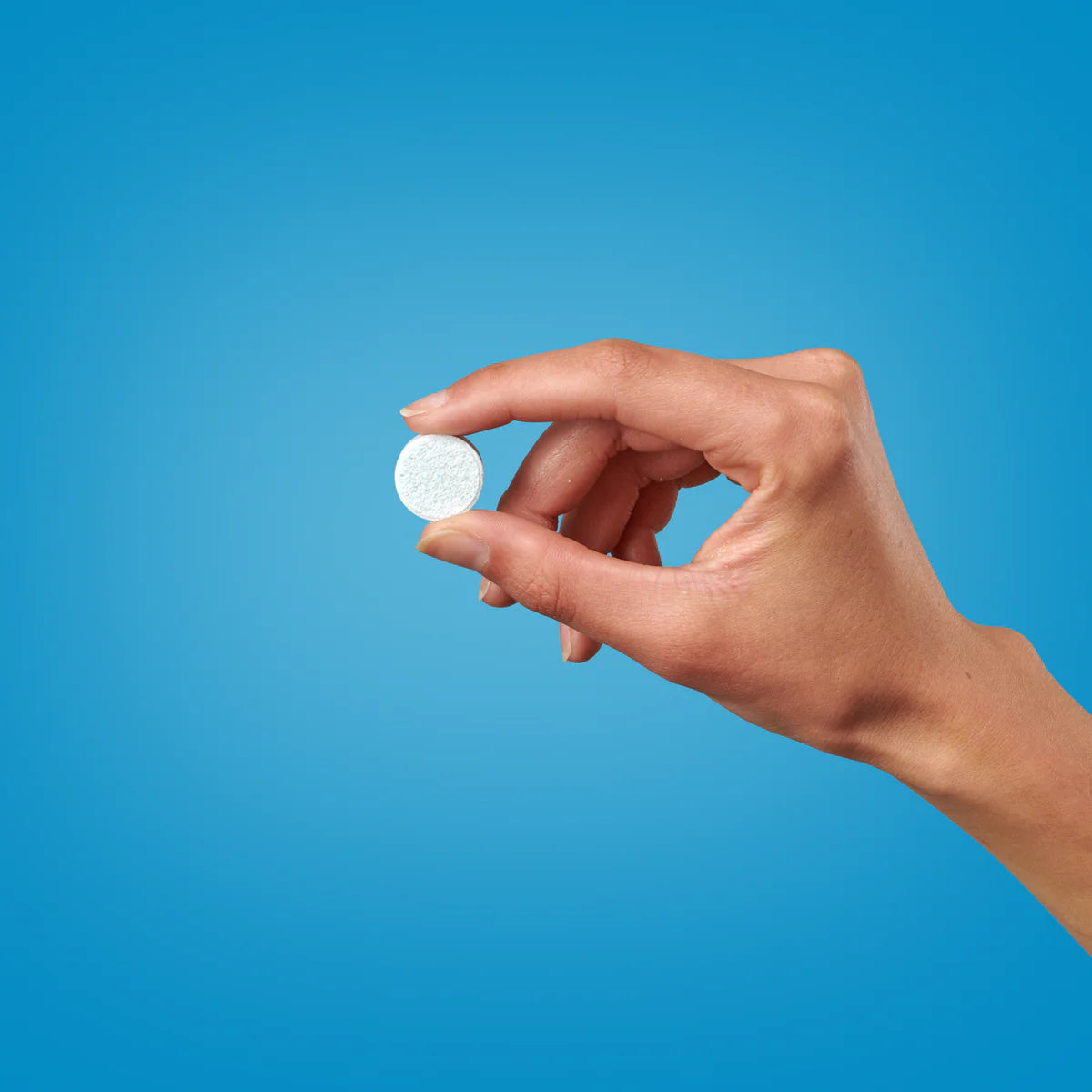 Eine Hand mit manikürten Nägeln hält eine runde weiße Tablette oder Pille zwischen Daumen und Zeigefinger vor einem schlichten blauen Hintergrund.