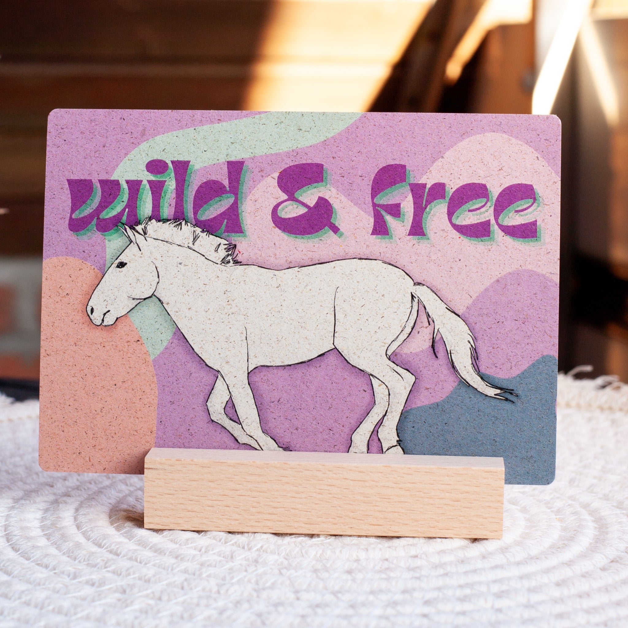 Eine Karte mit einem handgezeichneten weißen Pferd, das vor einem abstrakten Hintergrund in Pastellfarben läuft. Über dem Pferd steht in fetten, lila-grünen Buchstaben "wild & free" geschrieben. Die Karte wird auf einem Holzständer präsentiert.