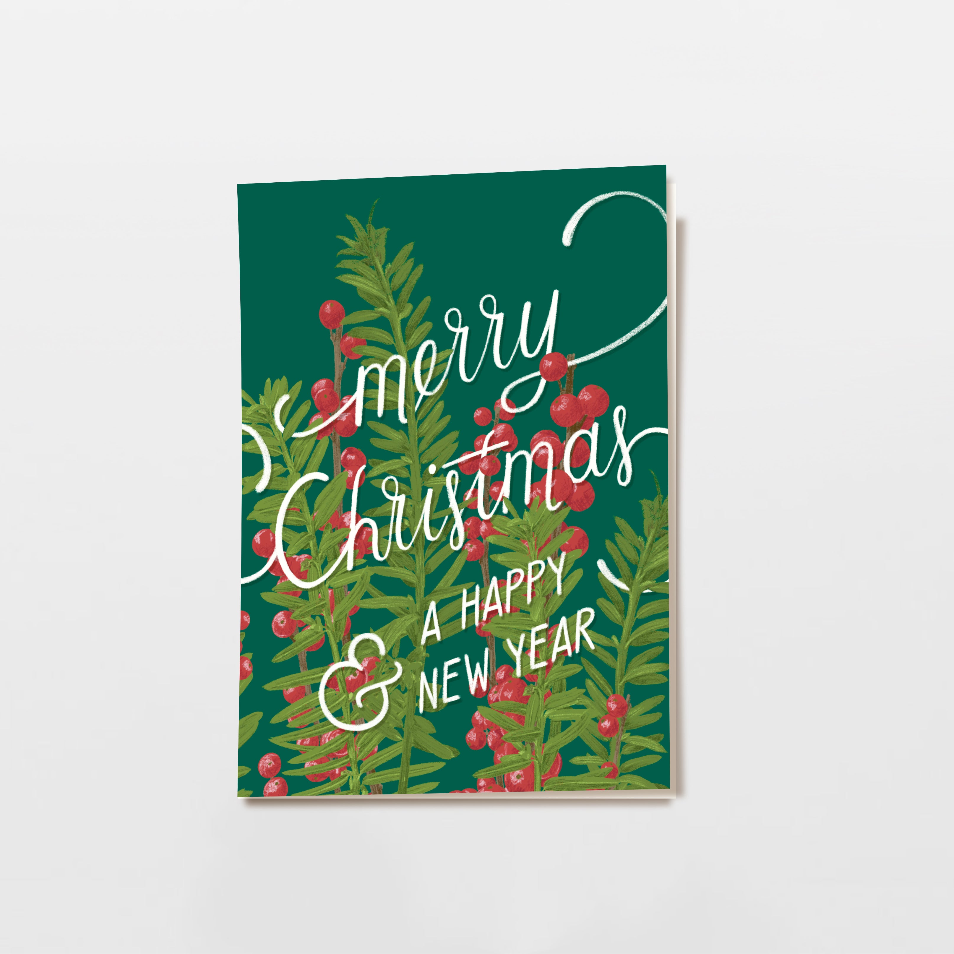 Grußkarte mit grünem Hintergrund und illustrierten Tannenzweigen mit roten Beeren. Der weiße handgeschriebene Text lautet: "Frohe Weihnachten und ein gutes neues Jahr". Die Karte steht aufrecht auf einer weißen Fläche.