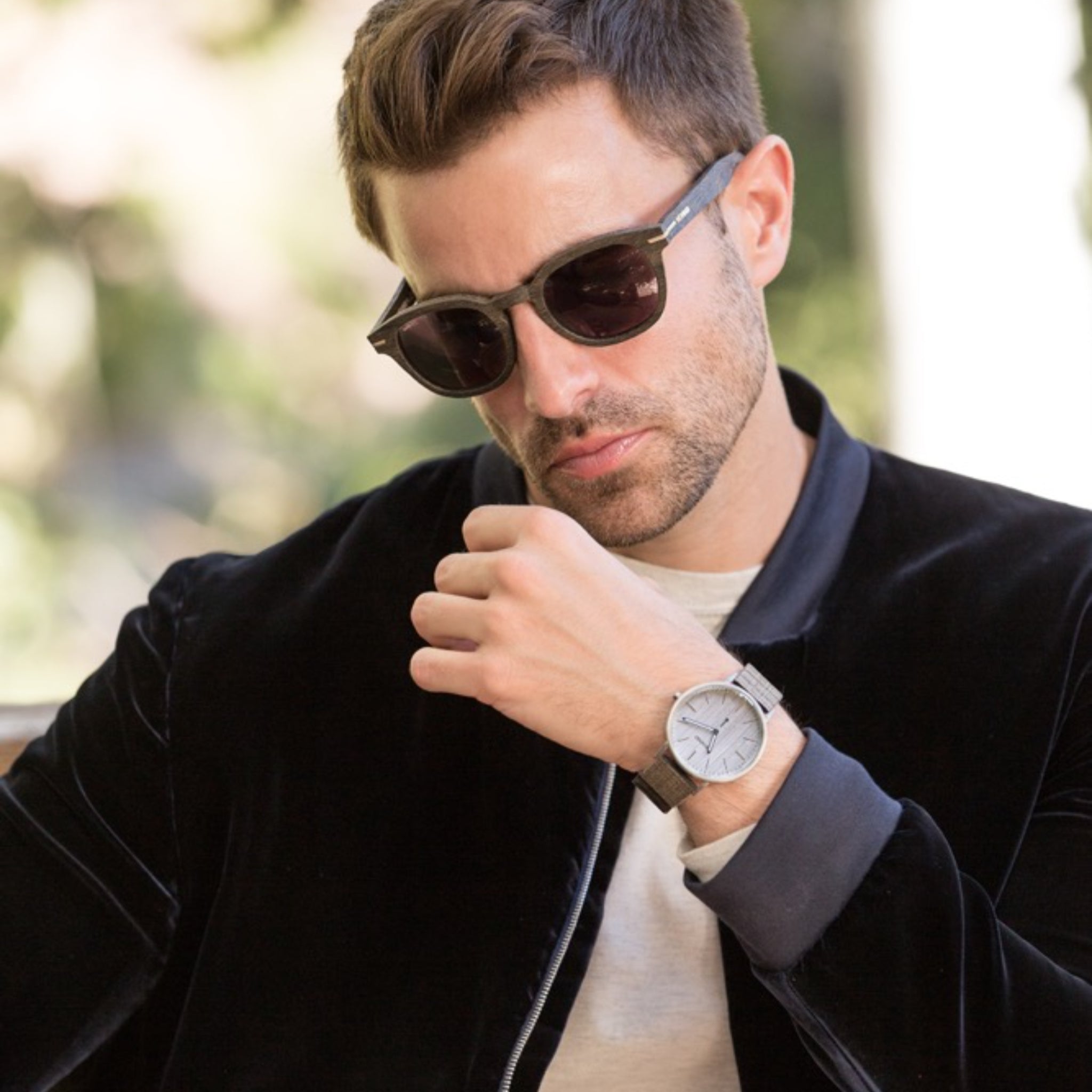Ein Mann mit Sonnenbrille und dunkler Jacke blickt nachdenklich nach unten und zeigt eine silberne Armbanduhr an seinem linken Handgelenk in einem Außenbereich.
