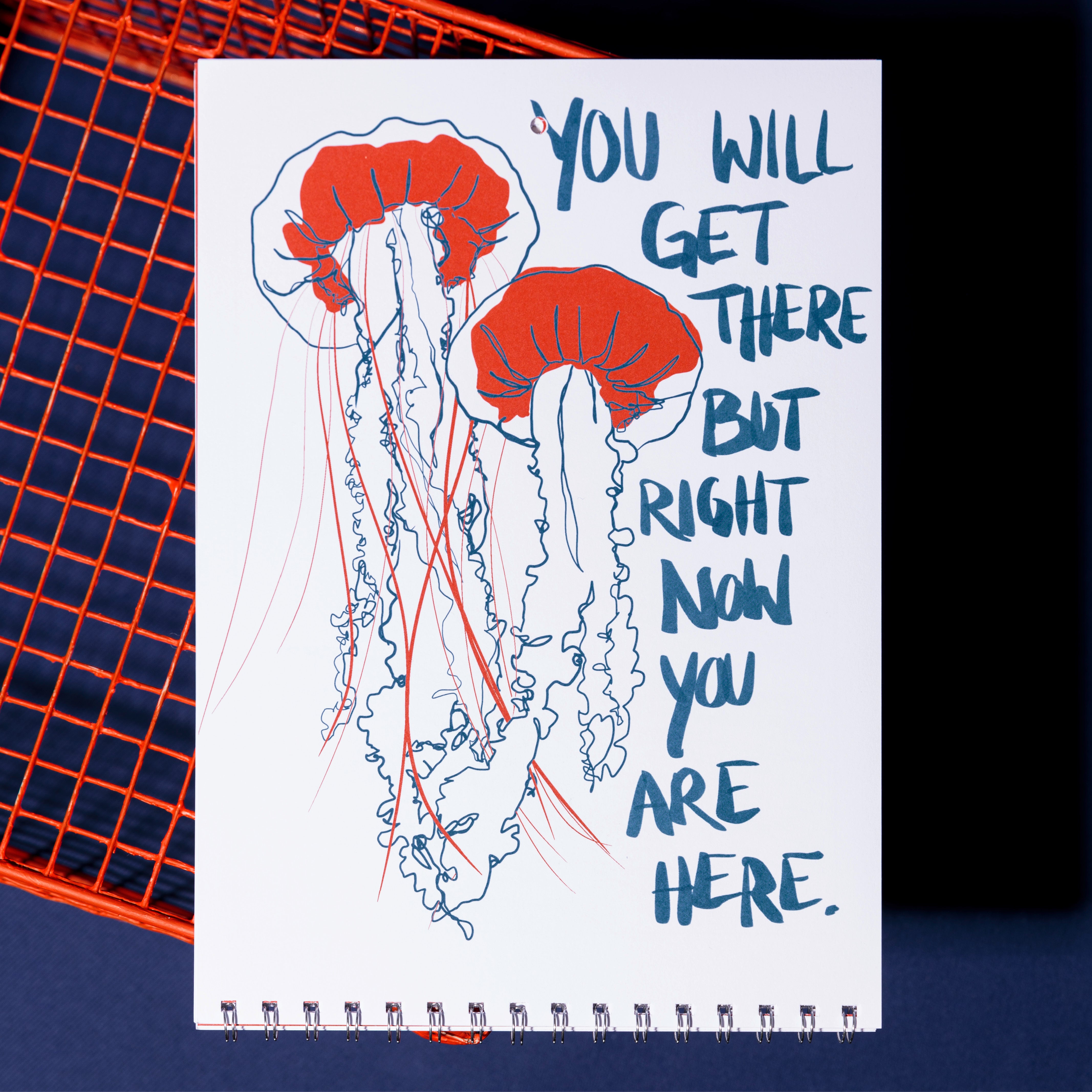 Eine Skizzenbuchseite zeigt zwei rote und blaue Quallen mit der handgeschriebenen Botschaft: Du wirst es schaffen, aber im Moment bist du hier. Das Skizzenbuch ruht auf einem orangefarbenen Drahtrahmen auf dunkelblauem Hintergrund.