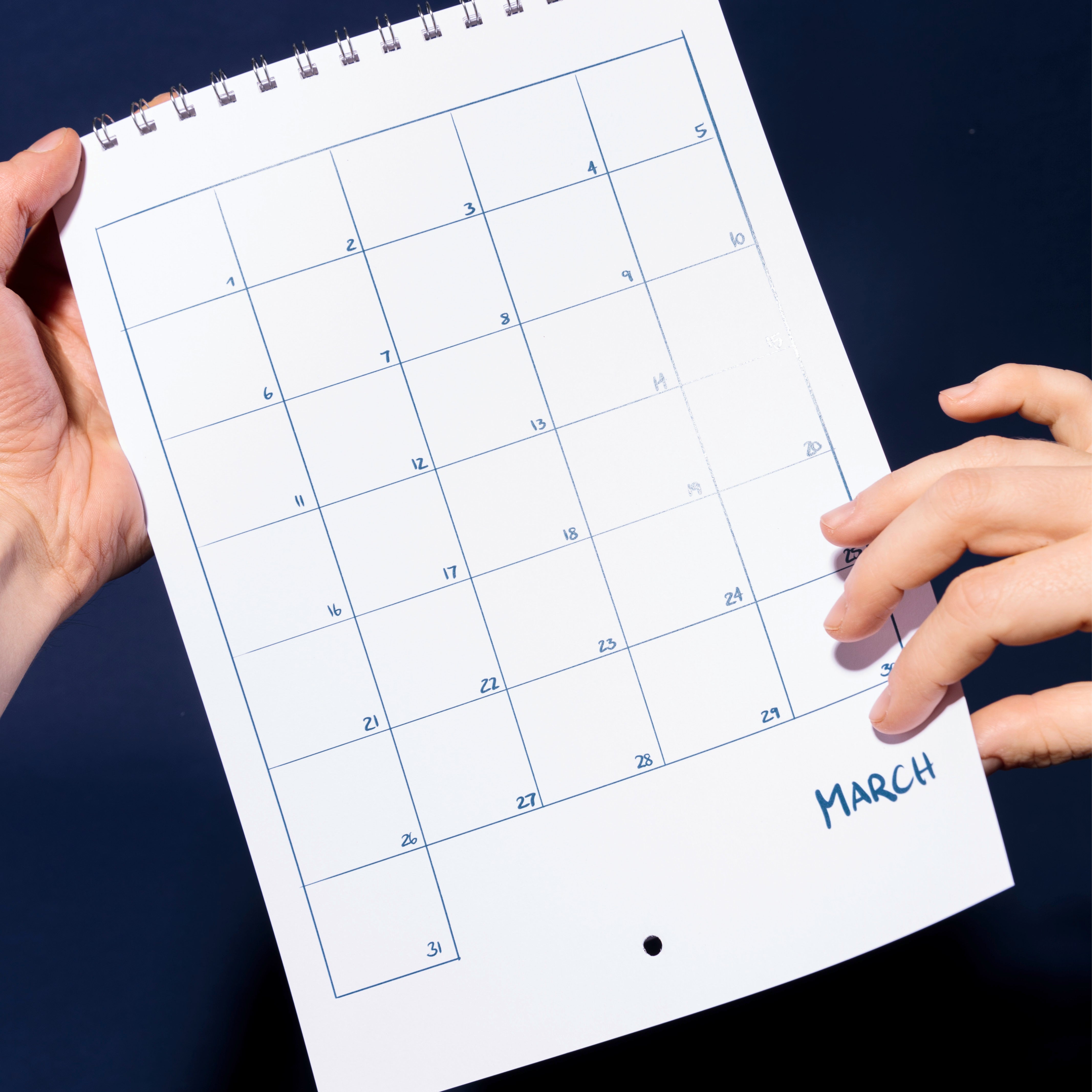 Eine Person hält einen spiralgebundenen Kalender mit dem Monat März in der Hand, dessen Datumsfelder leer sind und in dem keine Ereignisse oder Notizen eingetragen sind. Der Hintergrund ist dunkelblau.
