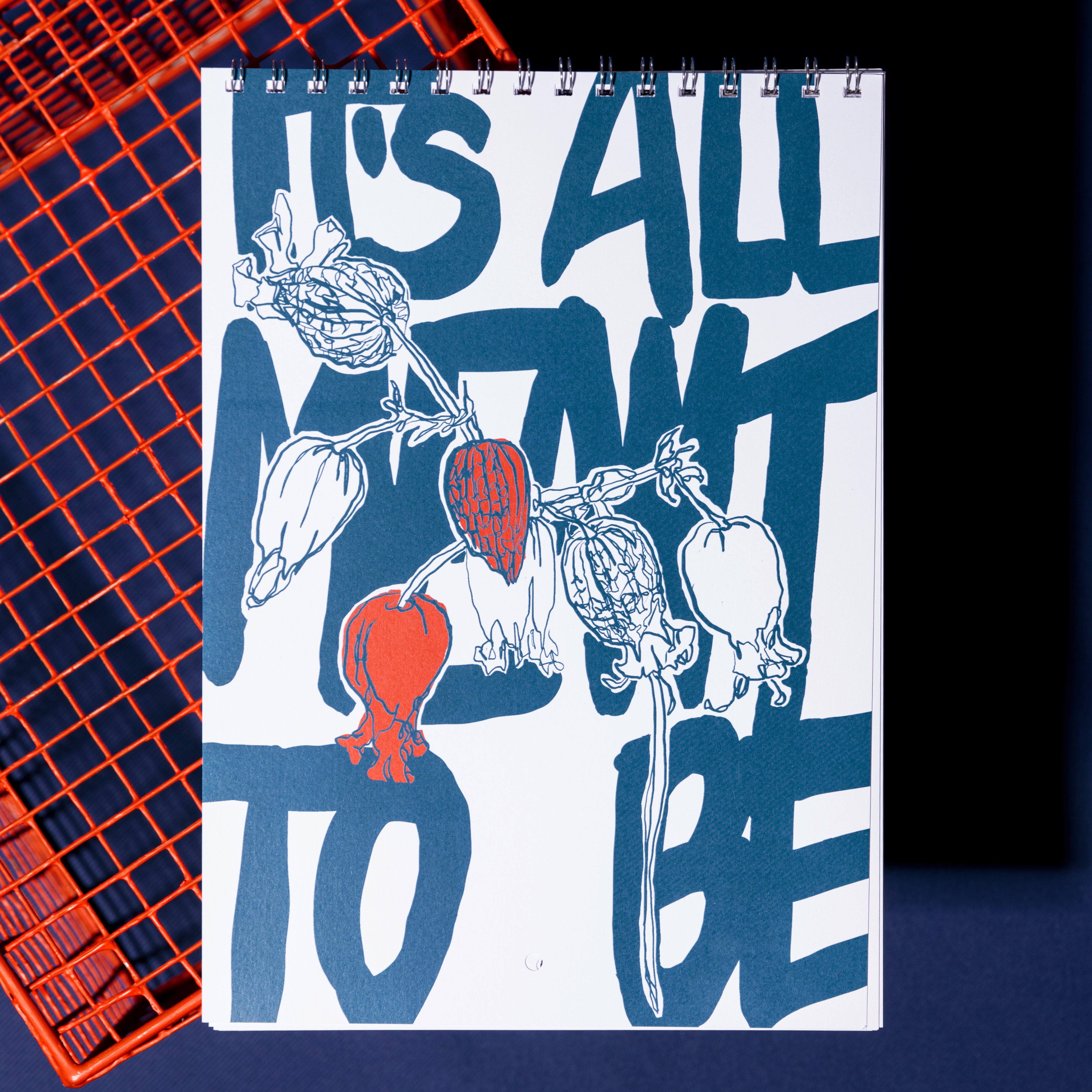 Das spiralgebundene Notizbuch zeigt den fetten blauen Text ITS ALL MEANT TO BE hinter einem illustrierten botanischen Design mit einer roten und mehreren umrandeten Beeren vor einem orangefarbenen Drahtgitterhintergrund.