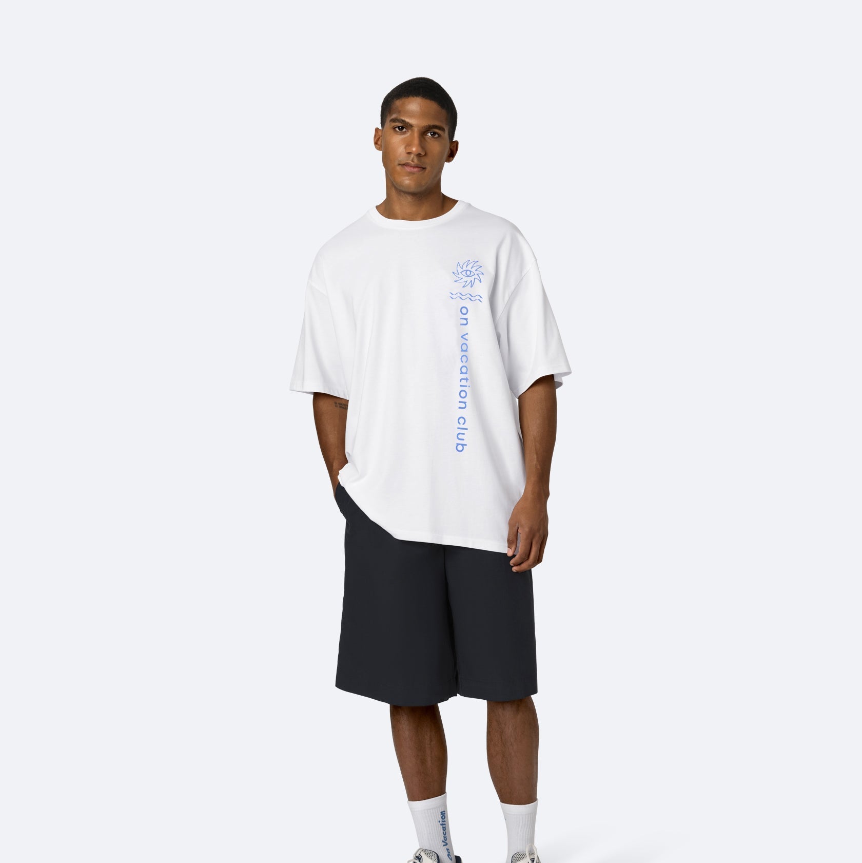 Ein Mann steht mit dem Gesicht nach vorne und trägt ein weißes übergroßes T-Shirt mit blauem Text, schwarze knielange Shorts, weiße Socken und weiße Turnschuhe. Er hat kurze Haare und einen neutralen Gesichtsausdruck vor einem schlichten hellen Hintergrund.