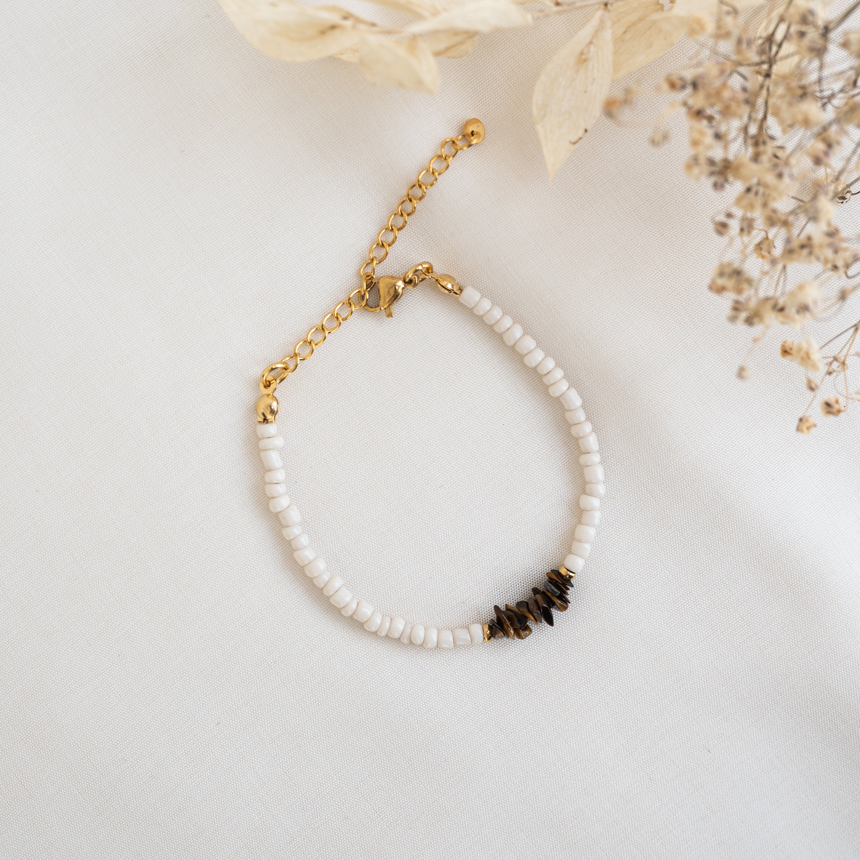 Ein zartes Armband mit kleinen weißen Perlen, einem goldenen Kettenverschluss und mehreren dunkelbraunen Steinen, die auf einem weißen Stoffhintergrund mit Trockenblumen in der Nähe angeordnet sind.