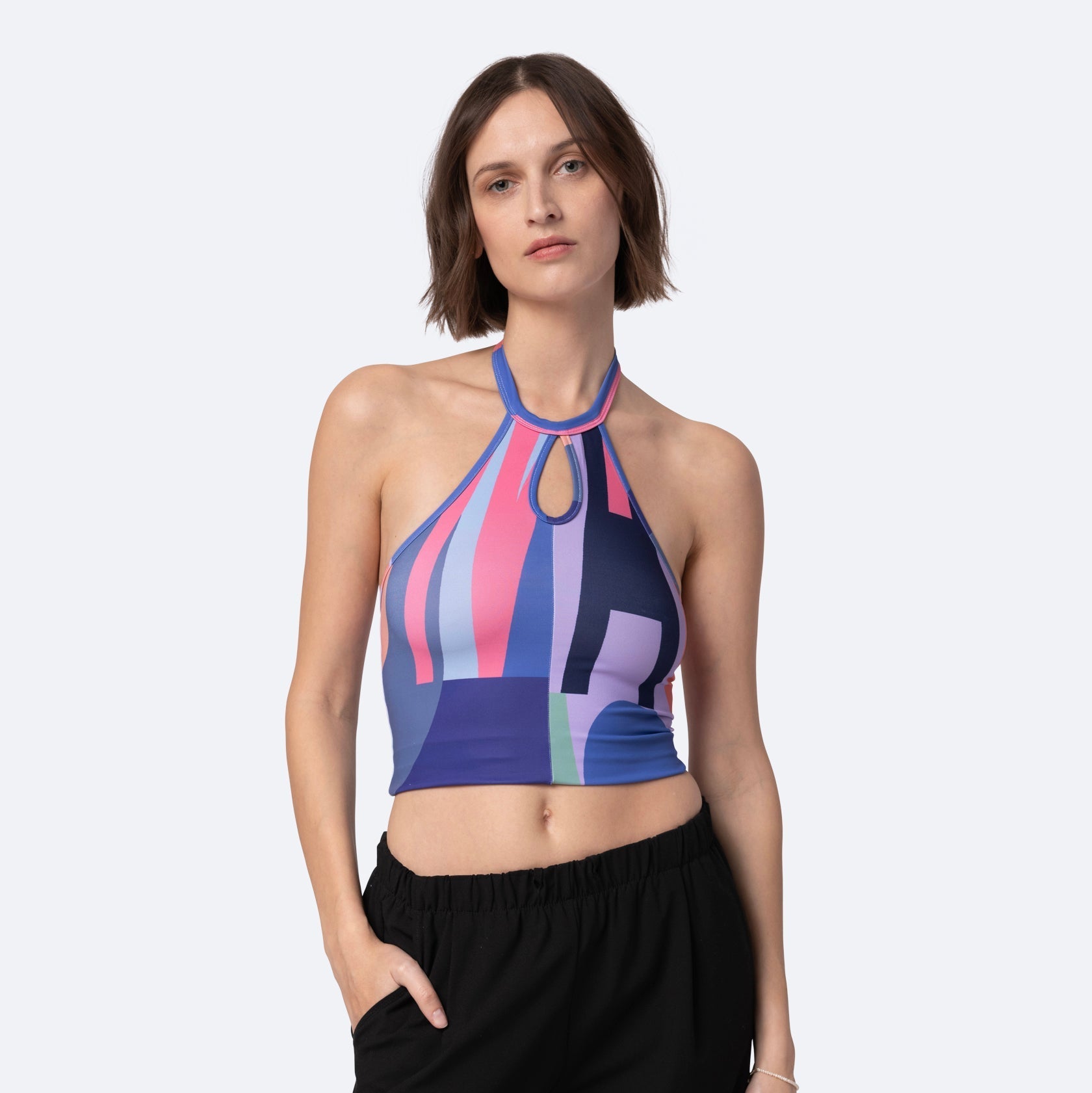 Eine Frau mit kurzen braunen Haaren trägt ein farbenfrohes Neckholder-Crop-Top mit geometrischen Mustern in Lila, Rosa und Blau sowie eine schwarze Hose. Sie steht vor einem schlichten hellen Hintergrund und hat eine Hand in der Tasche.