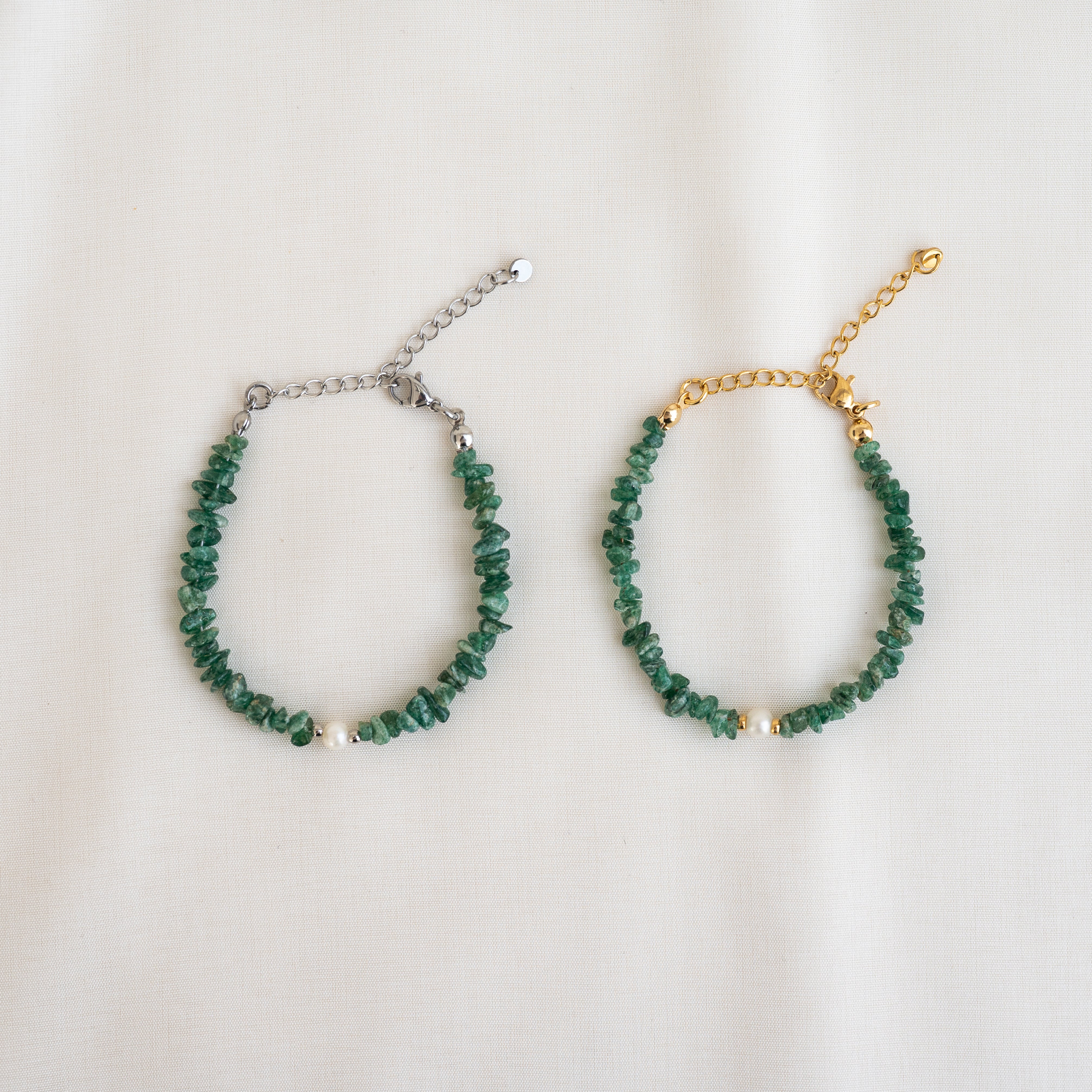Zwei grüne Perlenarmbänder mit verstellbaren Ketten, ein silberfarbenes und ein goldfarbenes, sind nebeneinander auf einem hellen Stoffhintergrund ausgestellt. In der Mitte jedes Armbands befindet sich eine einzelne weiße Perle.