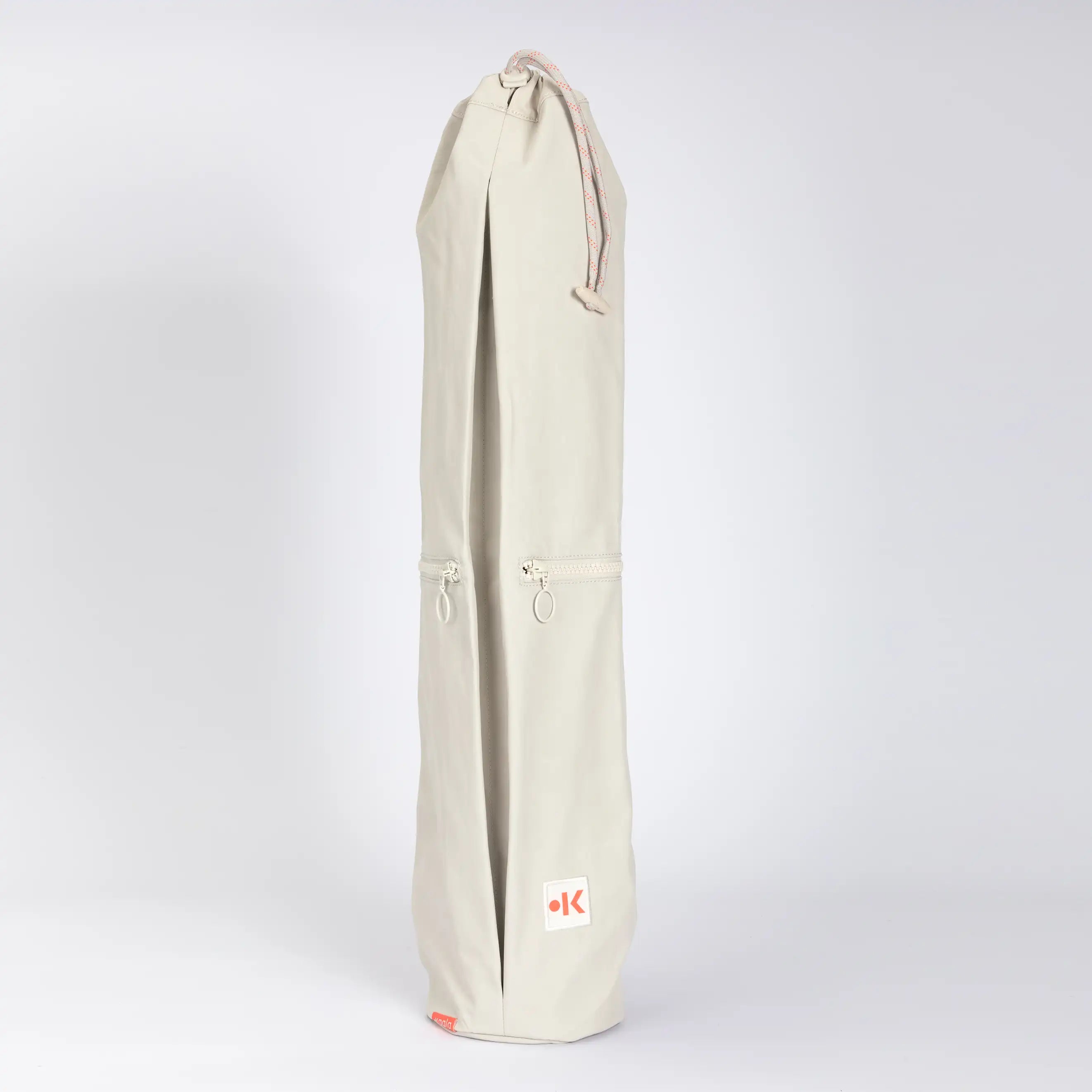 Beige Tasche mit Kordelzug und zwei Reißverschlusstaschen, aufrecht stehend vor grauem Hintergrund.