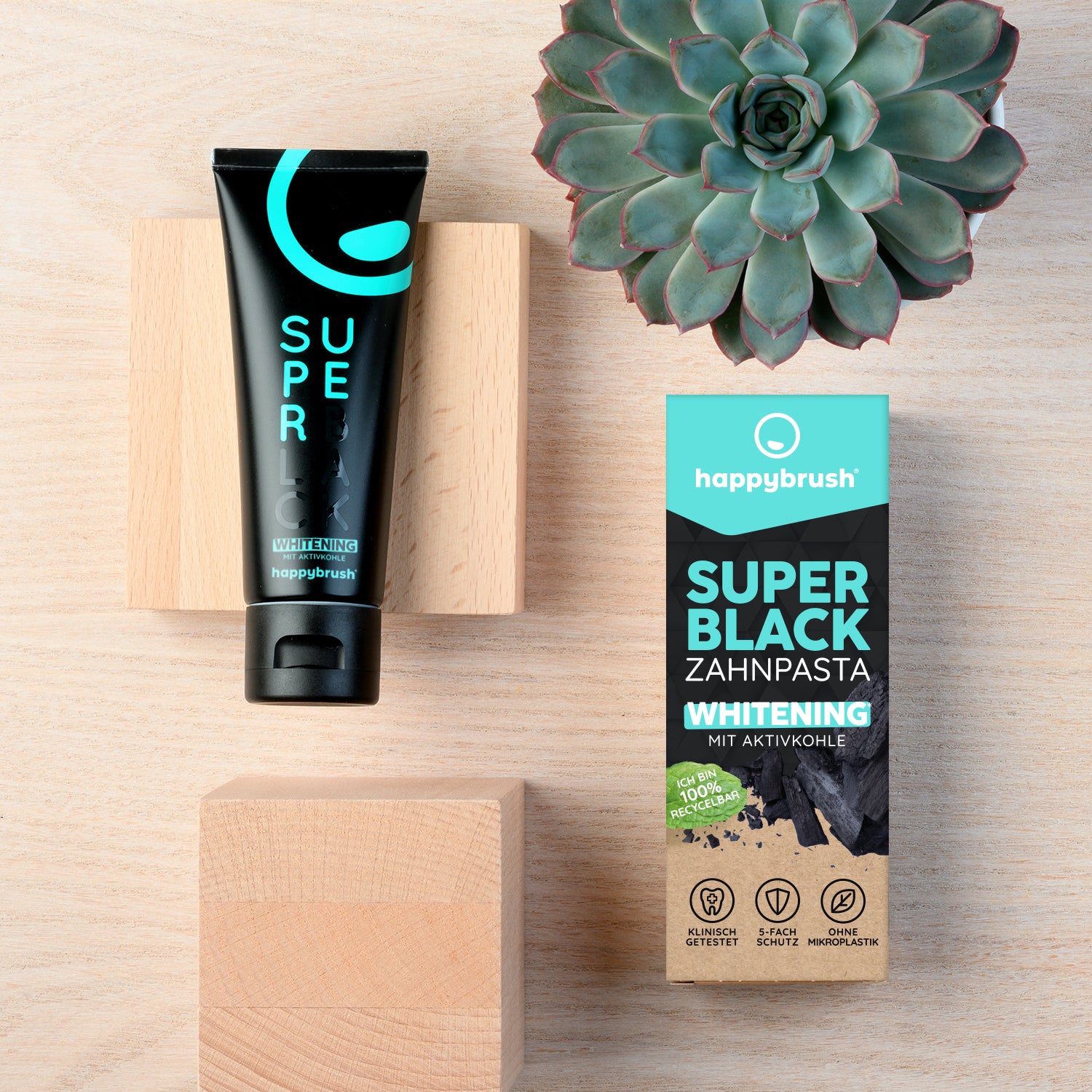 Schwarze Zahnpastatube und Verpackung von happybrush auf einer Holzoberfläche neben einer grünen Sukkulente.