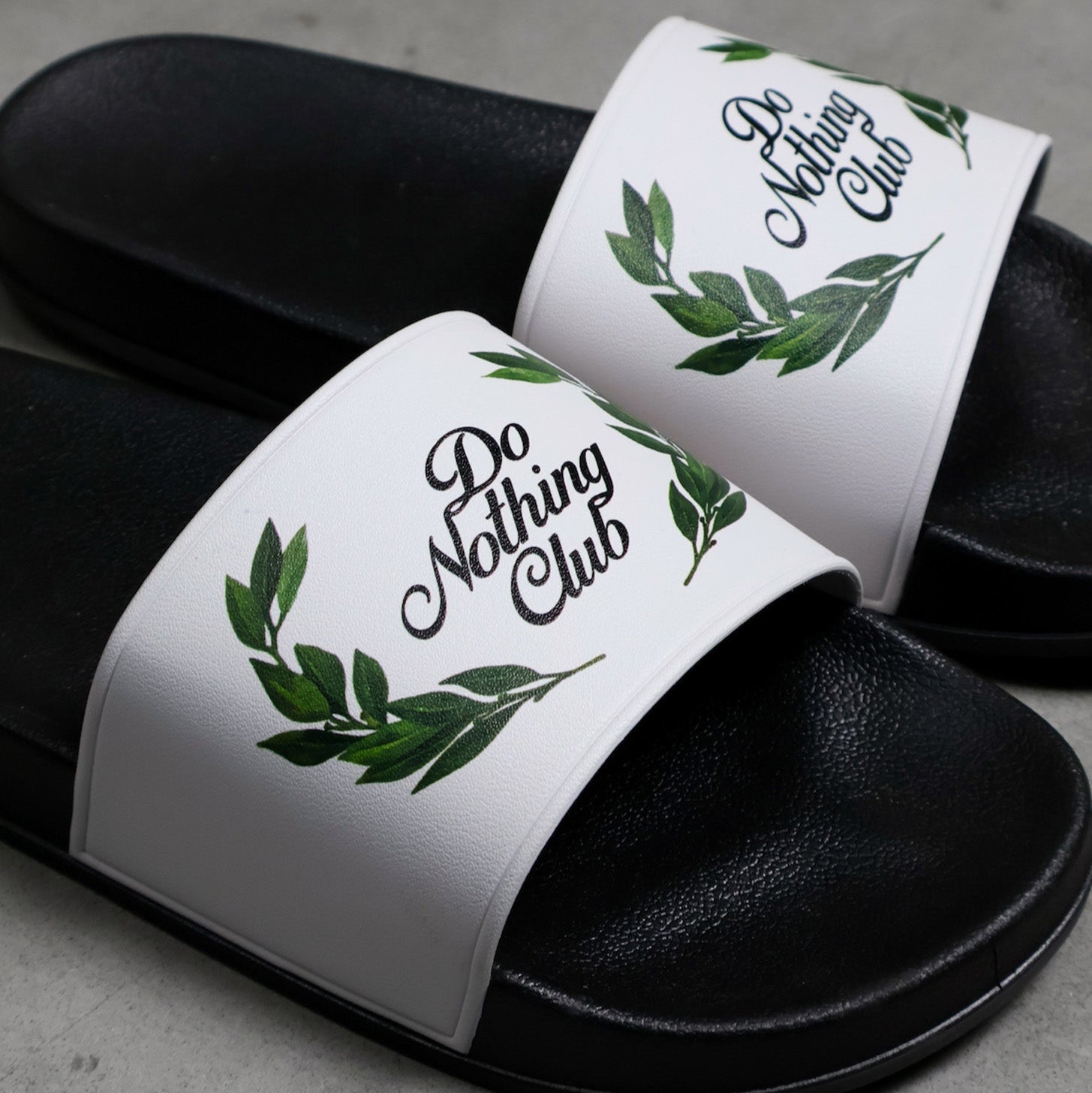 Ein Paar schwarze Slide-Sandalen mit weißen Riemen, die mit grünen Blattmustern und der Aufschrift Do Nothing Club bedruckt sind. Die Sandalen sind auf einer grauen Oberfläche platziert.