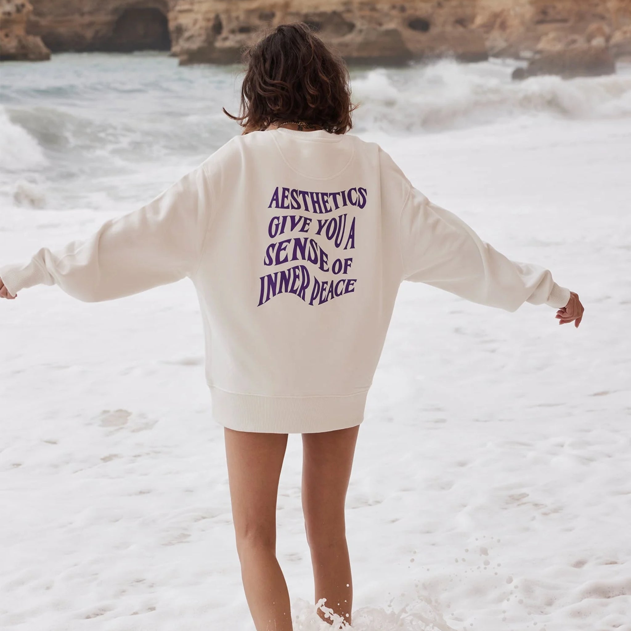 Person in white sweatshirt with text on back stands on a beach, facing the ocean.