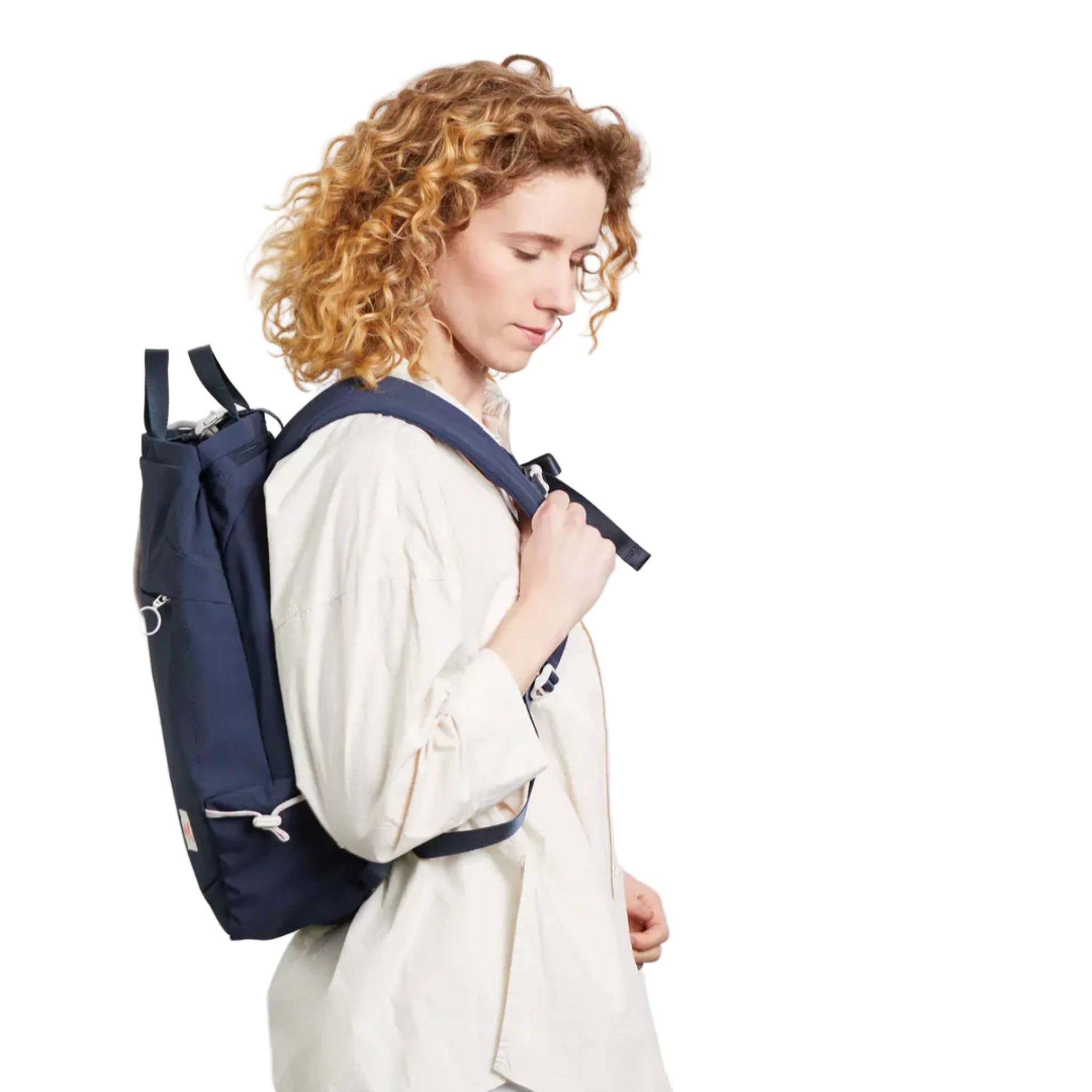 Eine Person mit lockigem blondem Haar, die ein lockeres weißes Hemd trägt, steht im Profil vor einem schlichten hellen Hintergrund und trägt einen marineblauen Rucksack über einer Schulter.